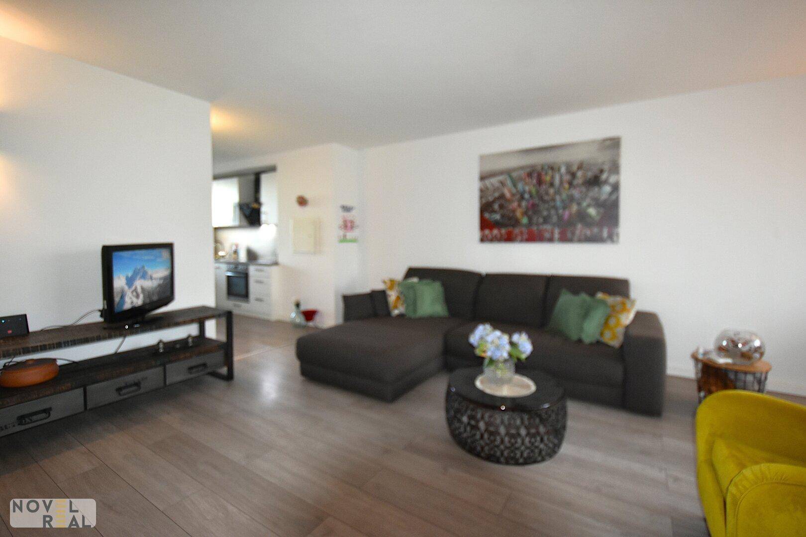 Komfortables Wohnzimmer mit dunklem Sofa, Fernseher und Blick auf die angrenzende Küche.
