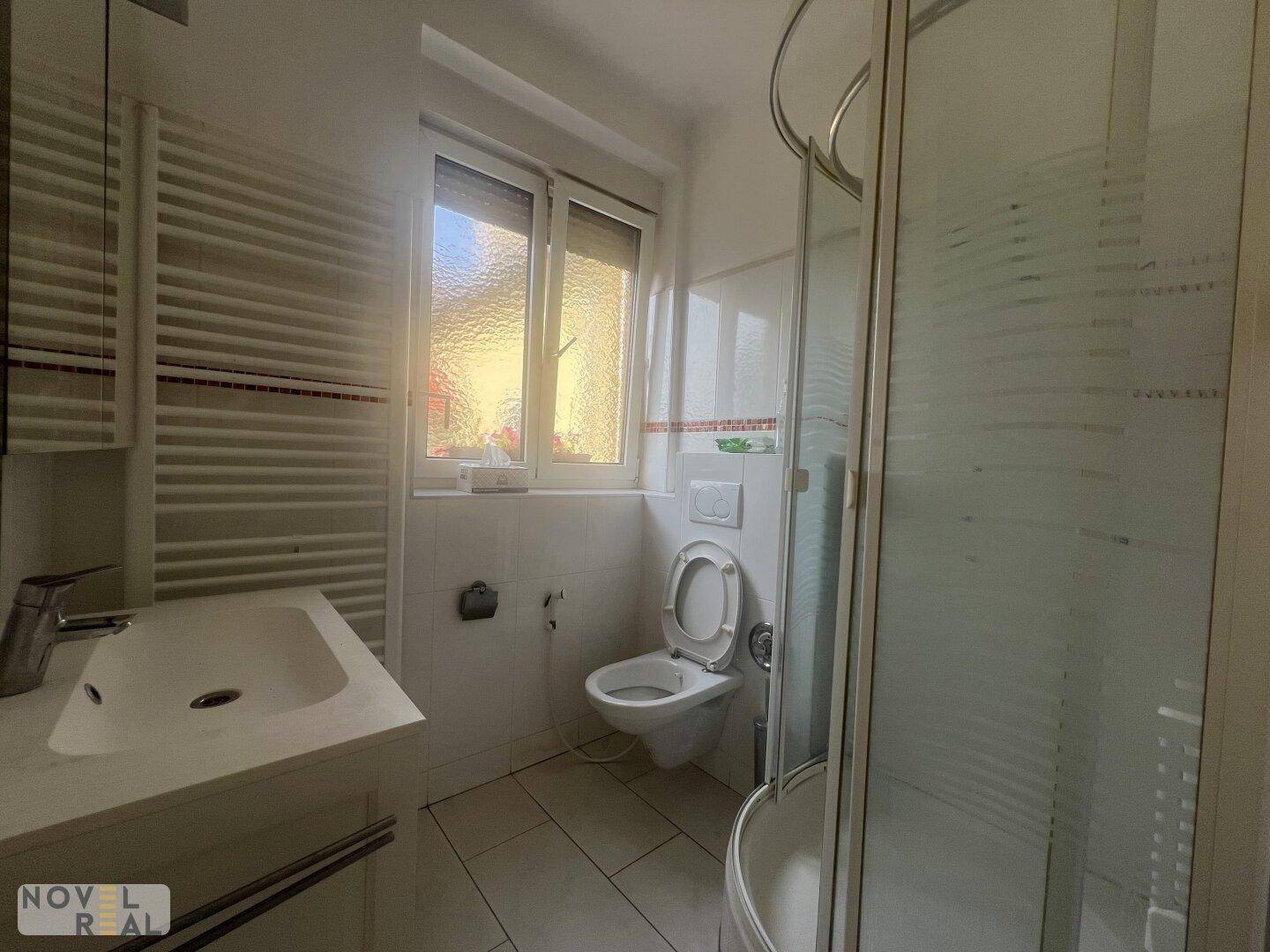 Helles Badezimmer mit Fenster, Toilette, Waschbecken und Duschkabine, modern gefliest.