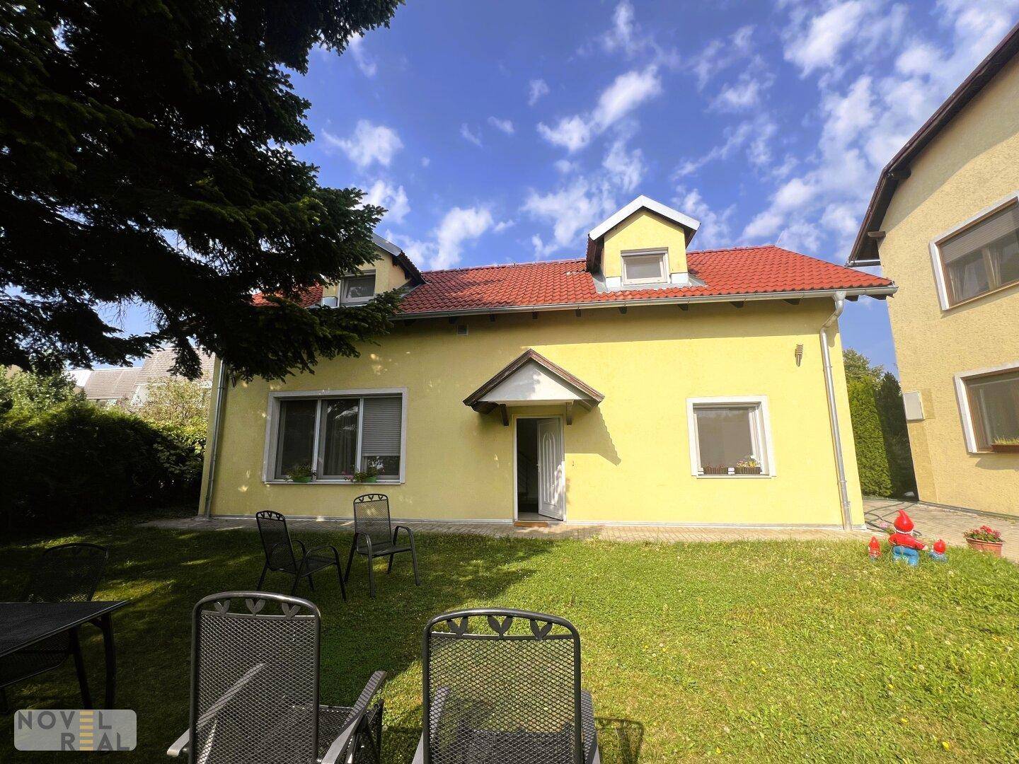 Gartenansicht des Hauses mit Rasenfläche und einfacher Sitzgruppe, umgeben von Bäumen und Sträuchern.