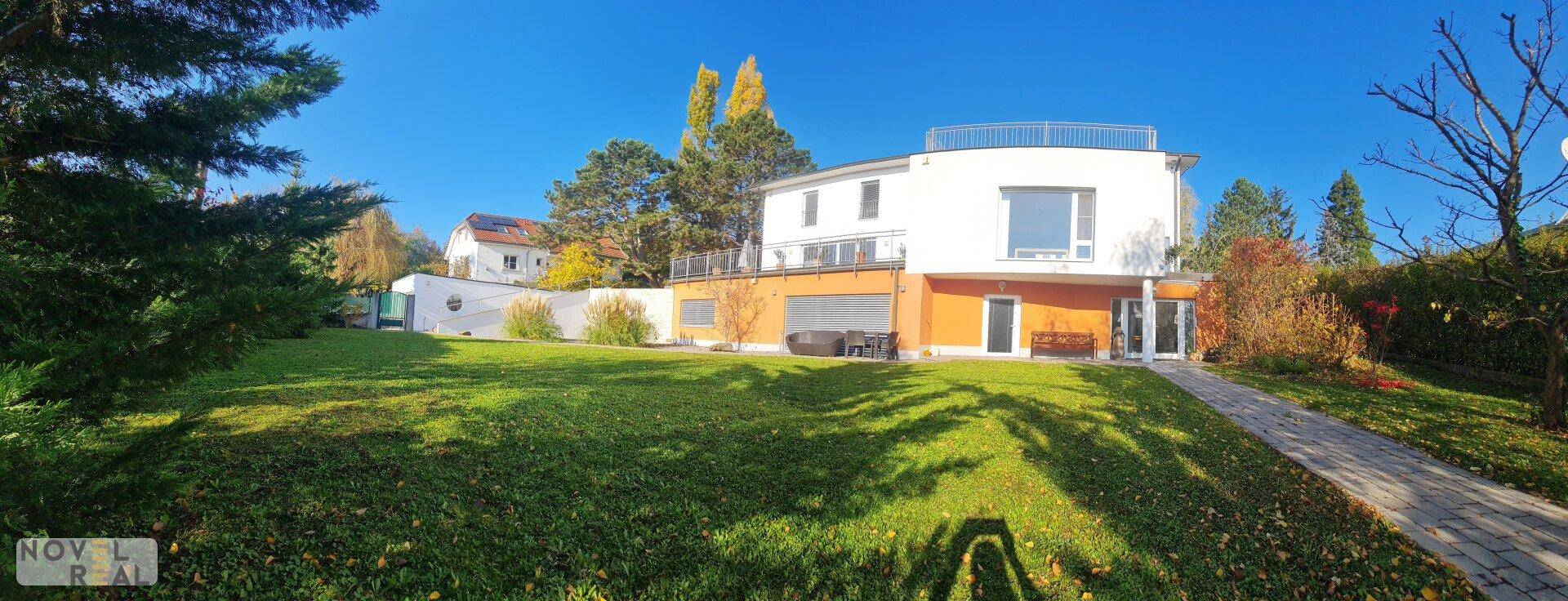 Weitläufiger Gartenbereich mit moderner Villa und umliegender Natur unter strahlend blauem Himmel.
