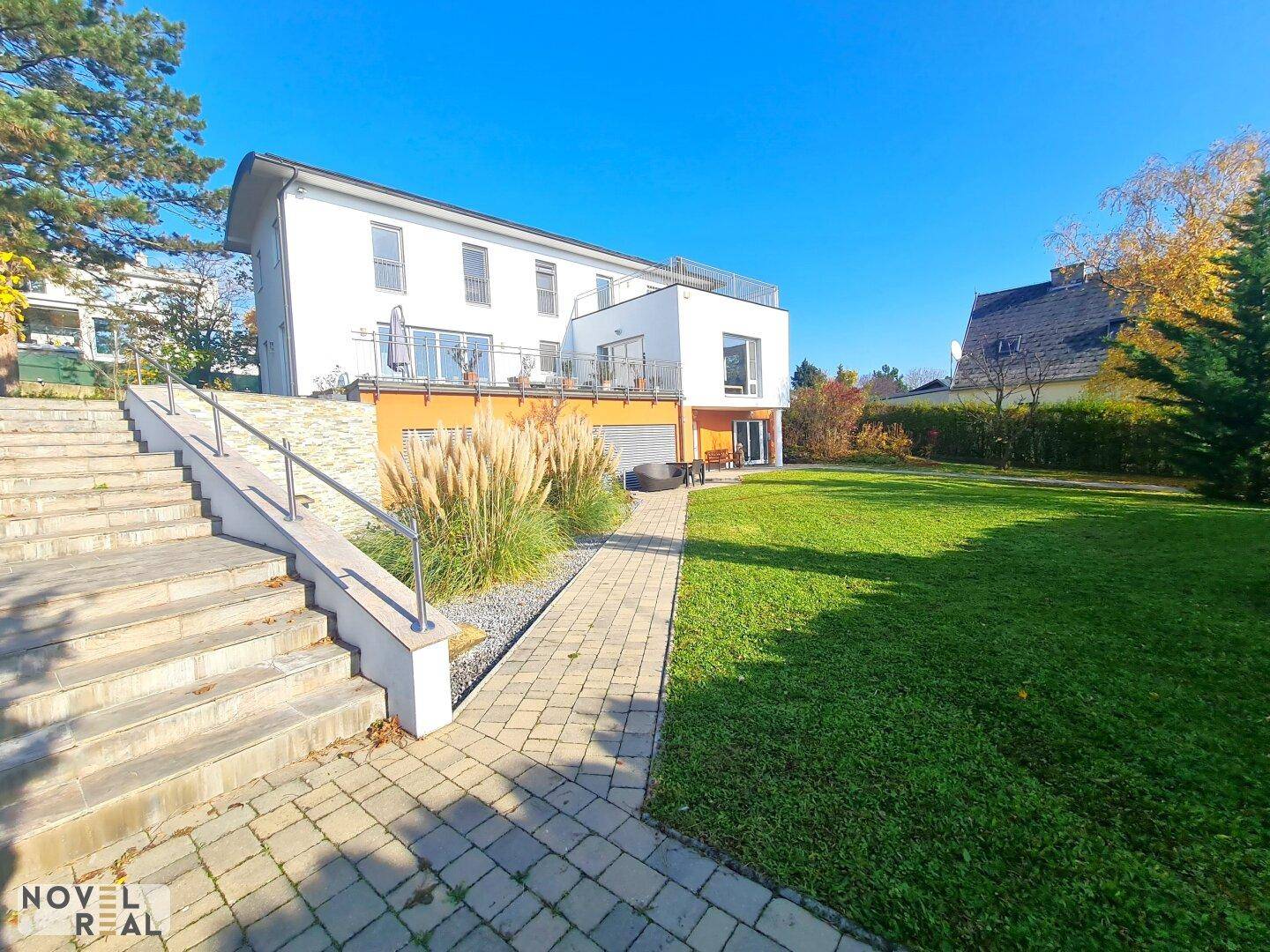 Modernes Haus mit großem Garten, gepflastertem Weg und Außentreppe unter blauem Himmel.