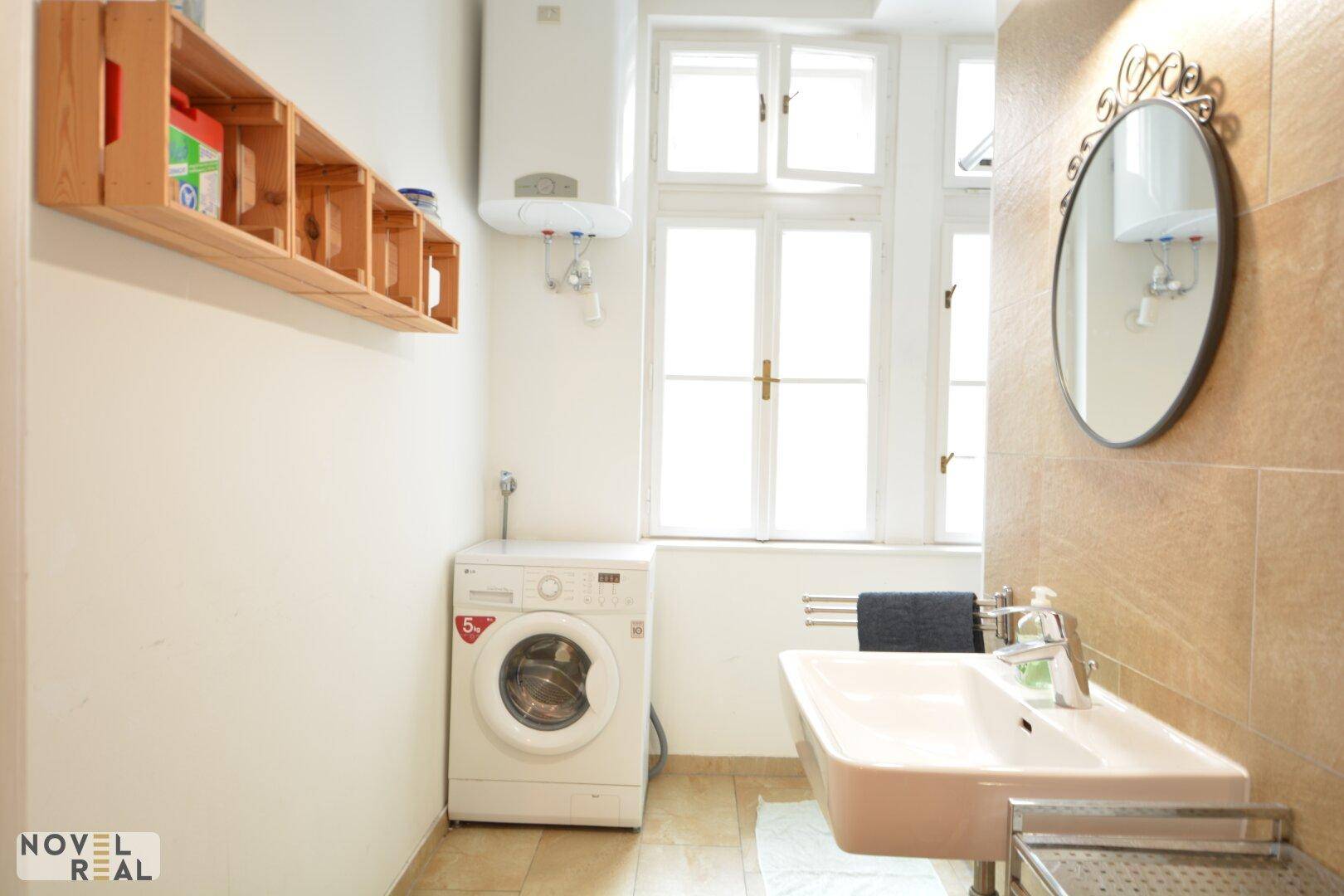 Helles Badezimmer mit Waschmaschine, Waschbecken, Fenster und beigefarbenen Fliesen.