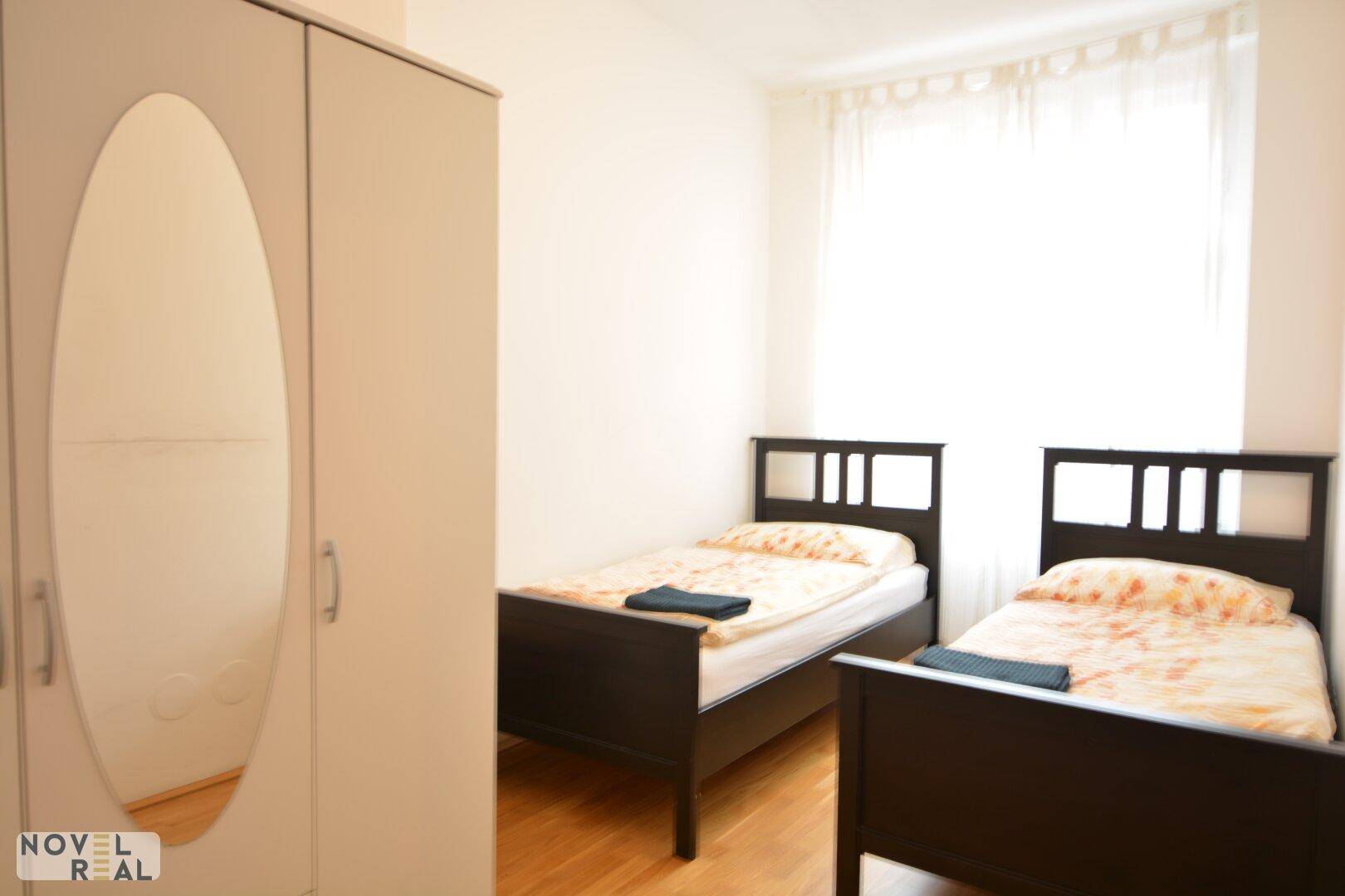 Helles Schlafzimmer mit zwei Einzelbetten, Kleiderschrank und Fenster mit Vorhängen.