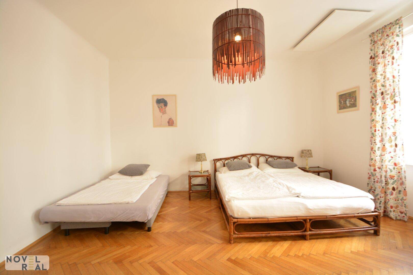 Helles Schlafzimmer mit Doppelbett, Einzelbett und Fischgrätparkettboden, gut beleuchtet durch ein Fenster.