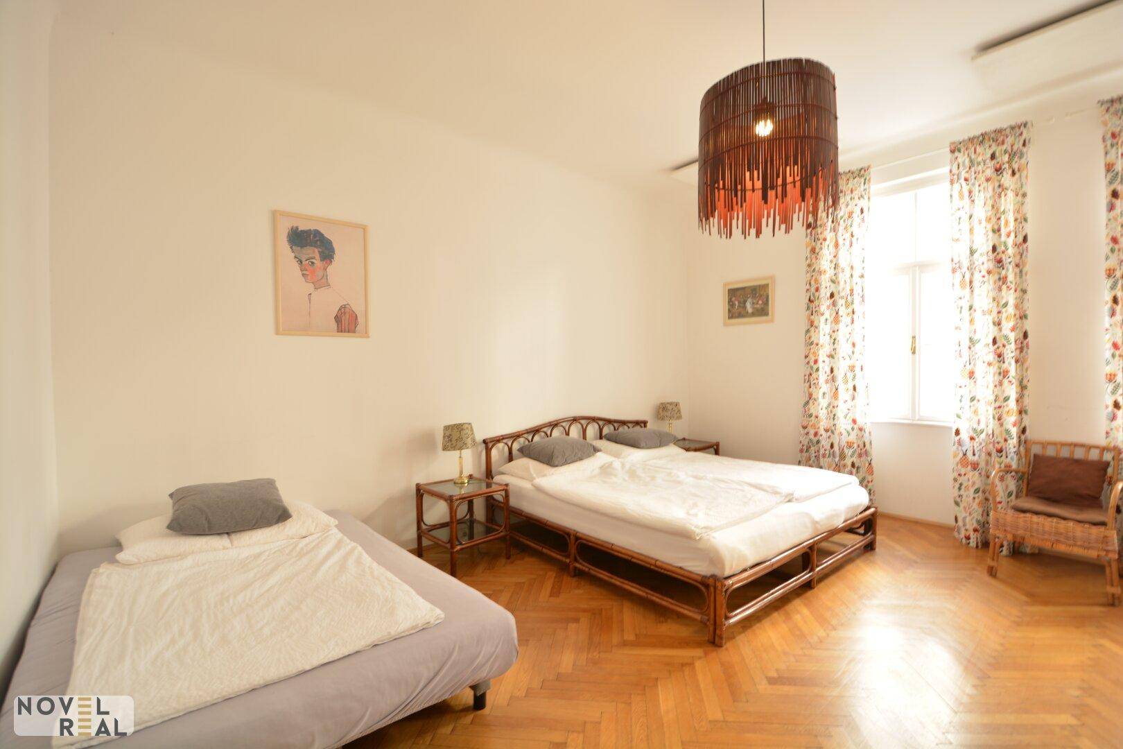 Geräumiges Schlafzimmer mit zwei Betten, Fischgrätparkett und einem großen Fenster mit bunten Vorhängen.