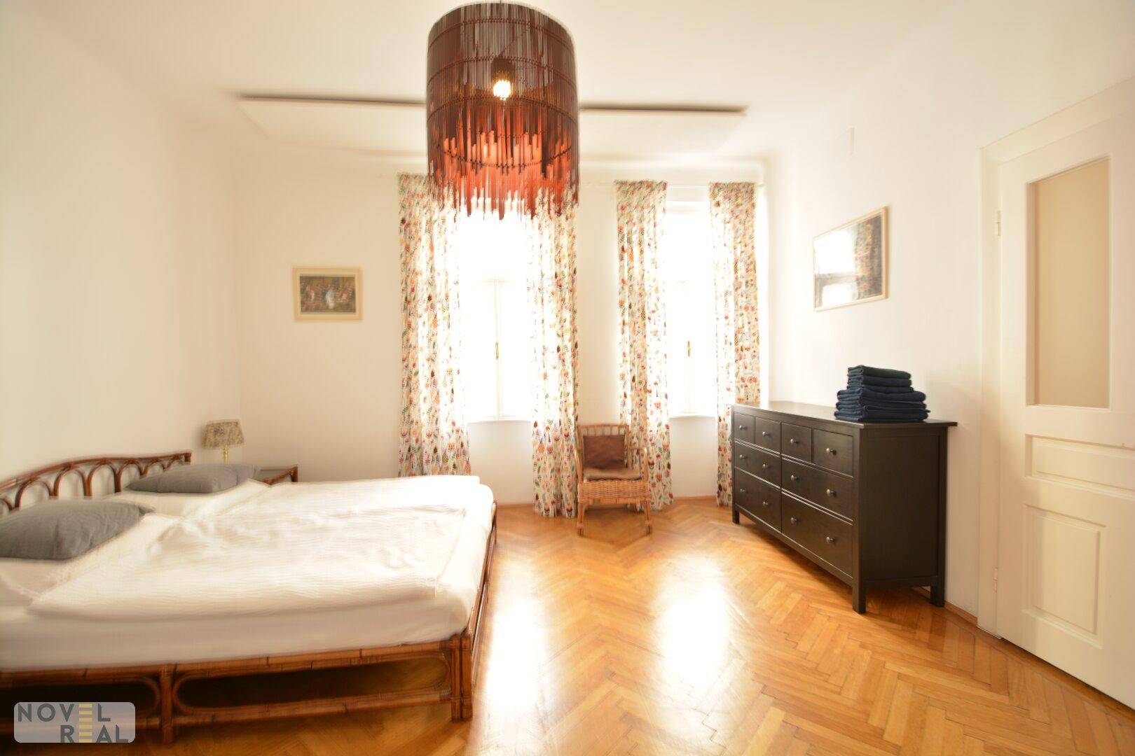 Helles Schlafzimmer mit Doppelbett, dunkler Kommode und zwei hohen Fenstern, die viel Tageslicht bieten.