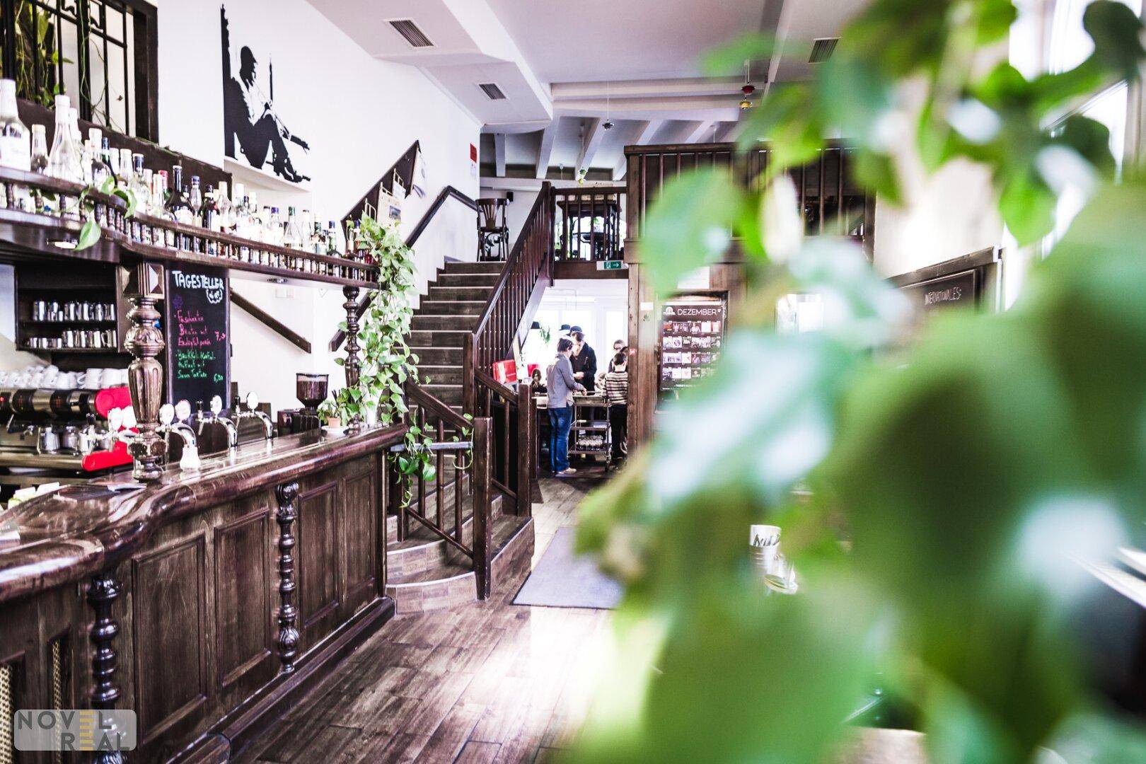 Rustikaler Restaurantbereich mit Holztheke, Treppe und vielen Pflanzen, hell beleuchtet.