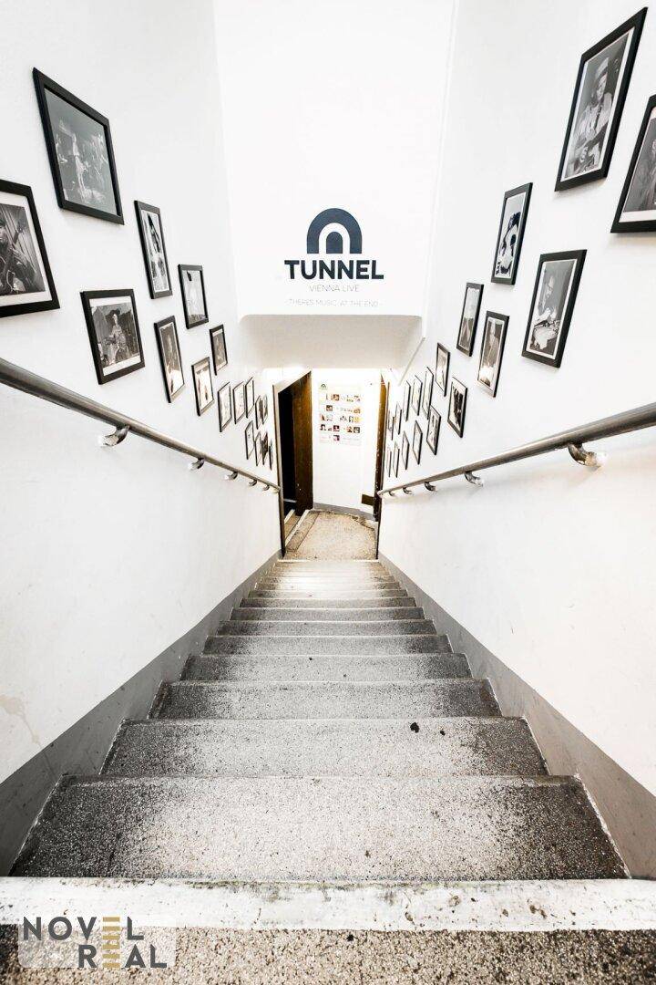 Heller Treppenaufgang mit weißen Wänden, gerahmten Schwarz-Weiß-Bildern und einem "TUNNEL"-Logo.
