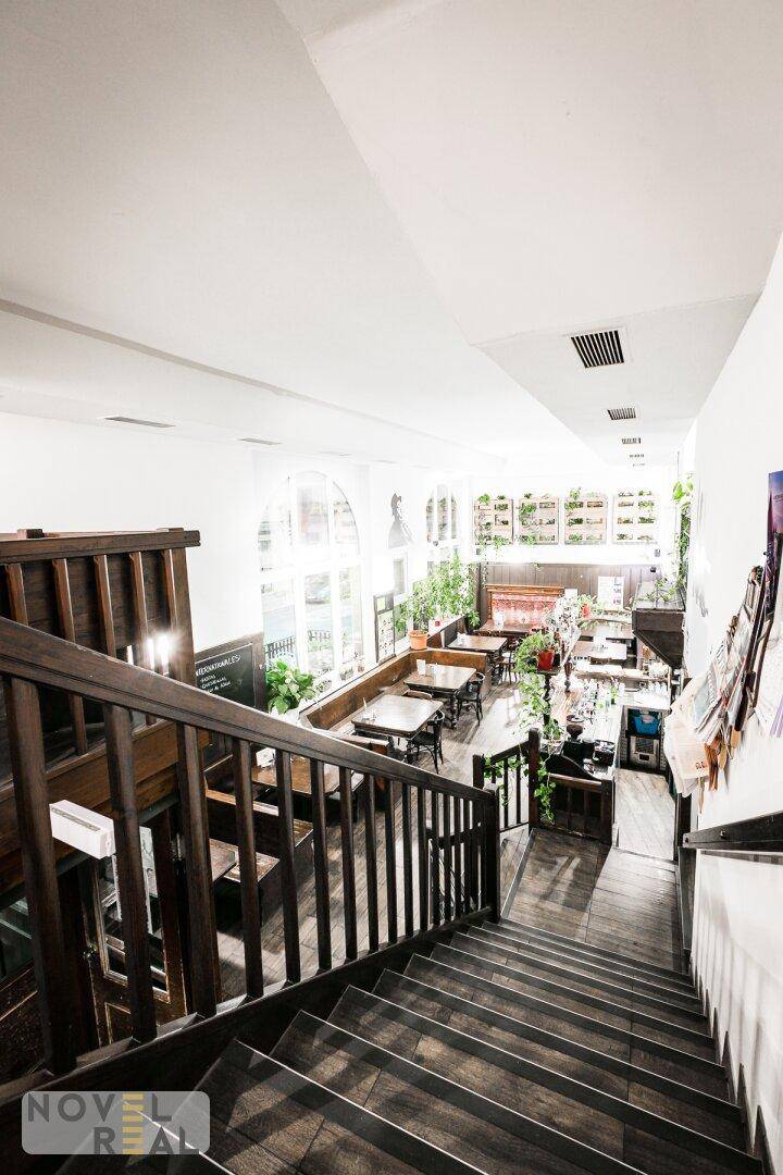 Blick von oben auf eine Holztreppe, die zu einem hellen Restaurantbereich mit großen Fenstern führt.
