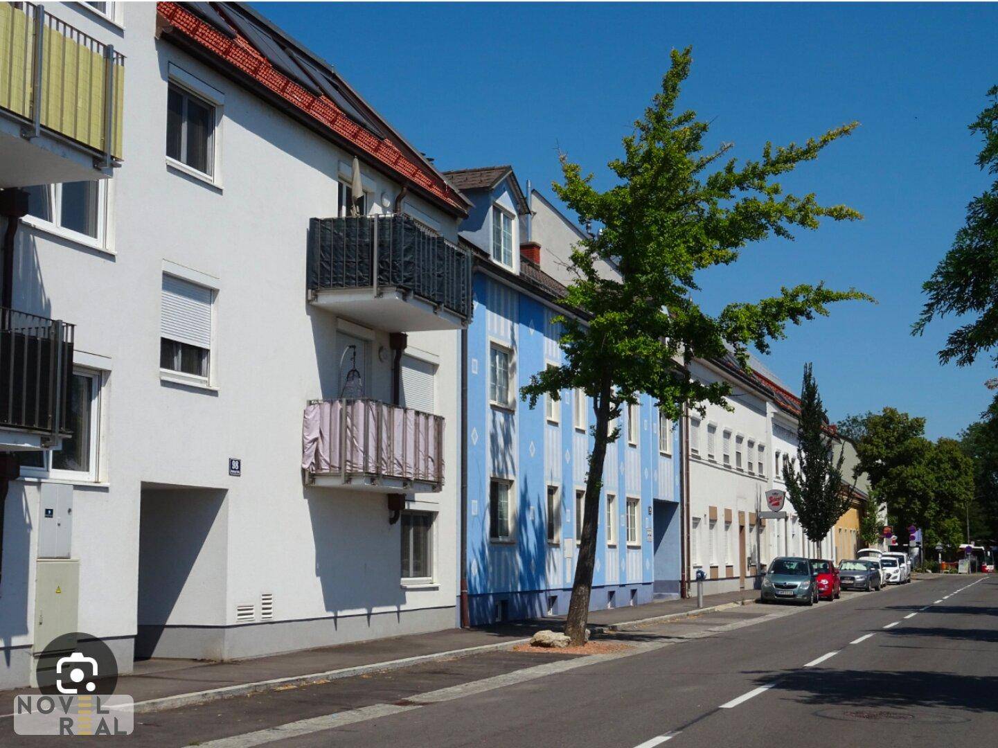 Wohnstraße mit Gebäuden in verschiedenen Farben, Balkonen und geparkten Autos unter blauem Himmel.