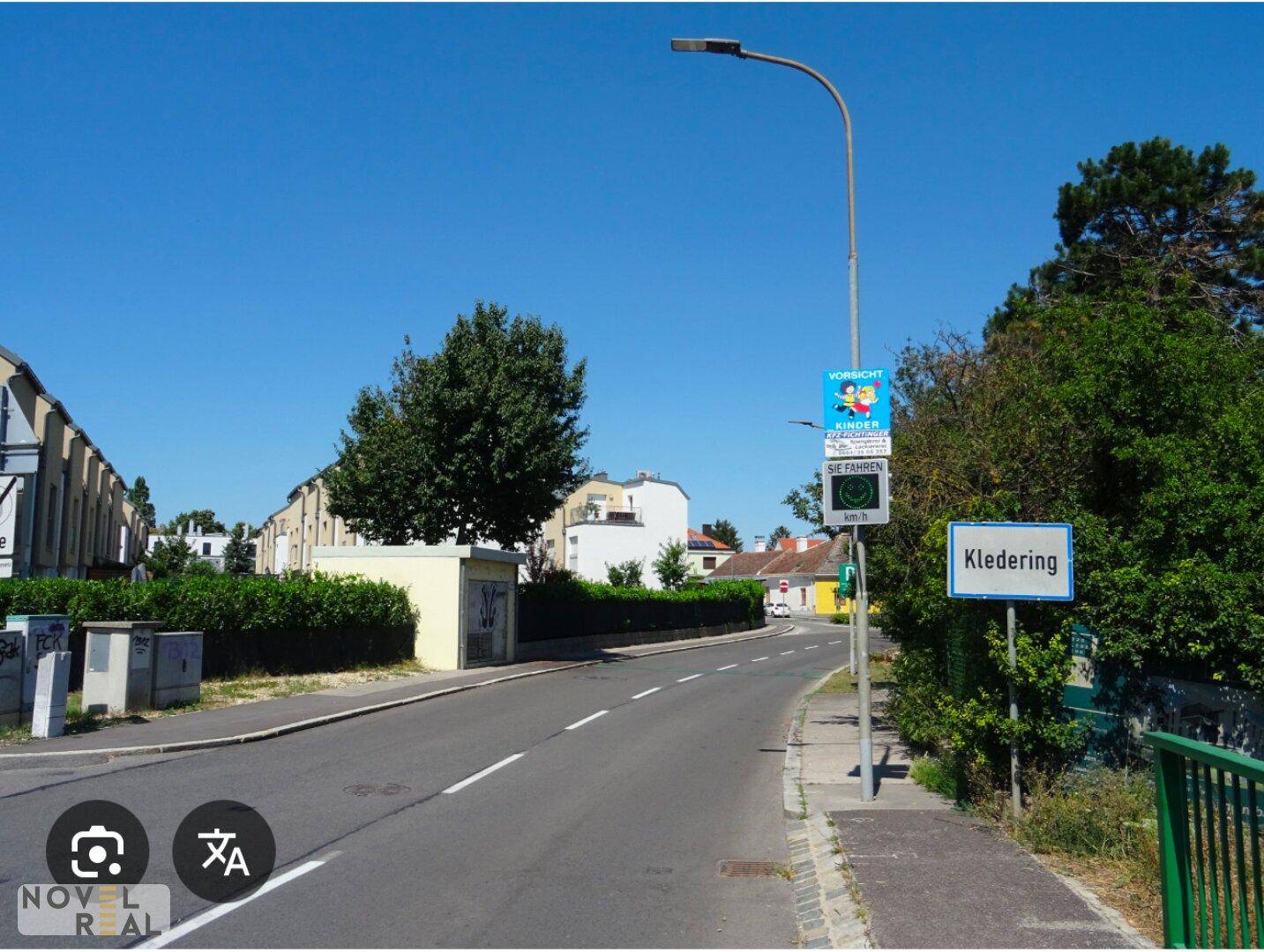 Einfahrt in die Ortschaft Kledering mit Straßenschild, Bäumen und Gebäuden unter blauem Himmel.