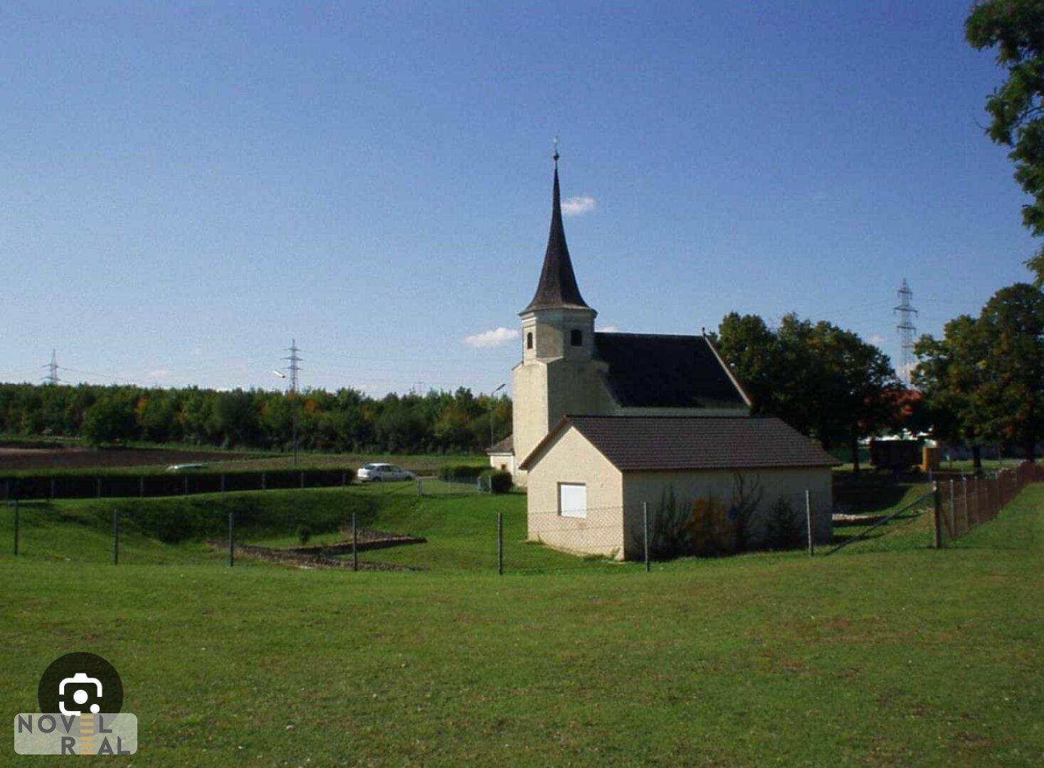 Kleine Kirche mit spitzem Turm, umgeben von weiten grünen Feldern und Bäumen.