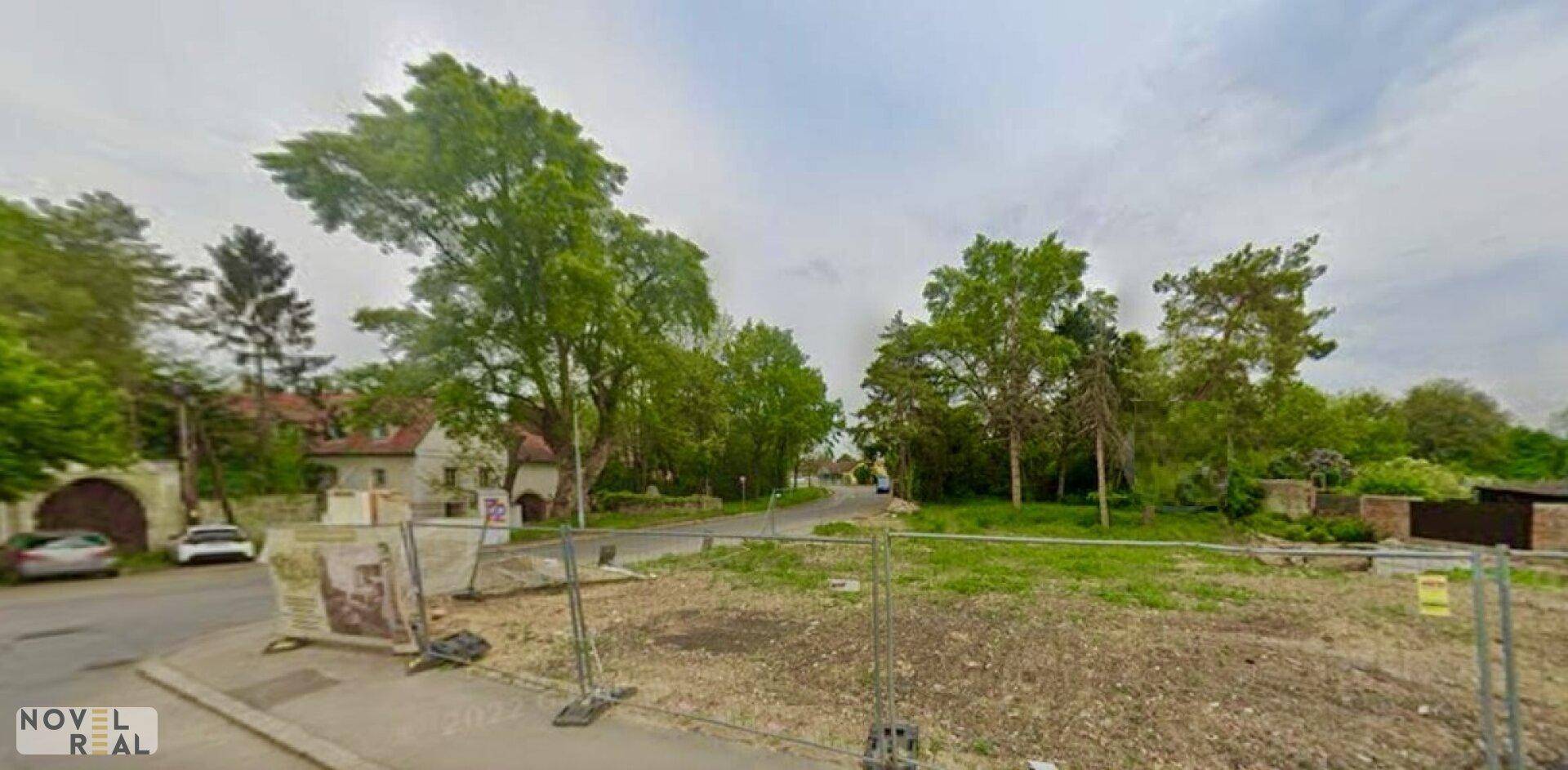 Leeres Grundstück mit Bauzaun, umgeben von Bäumen und einer Straße unter bewölktem Himmel.