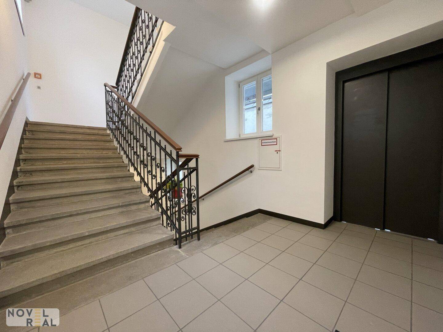Helles Treppenhaus mit Fliesenboden, schmiedeeisernem Geländer und modernem Aufzug.