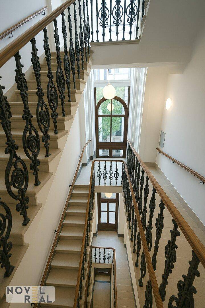 Historisches Treppenhaus mit schmiedeeisernem Geländer, Holzhandlauf und hohen Fenstern.