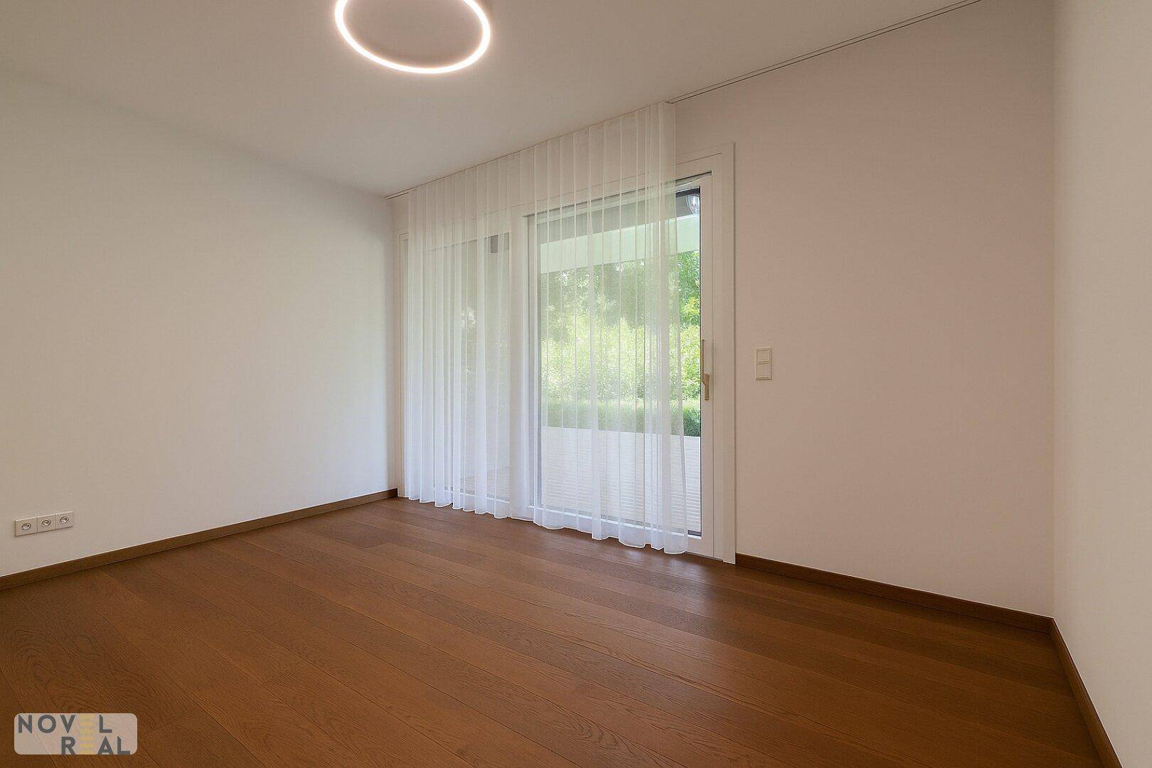 Leeres Zimmer mit hellem Holzfußboden und großer Glastür, die viel Licht hereinlässt.
