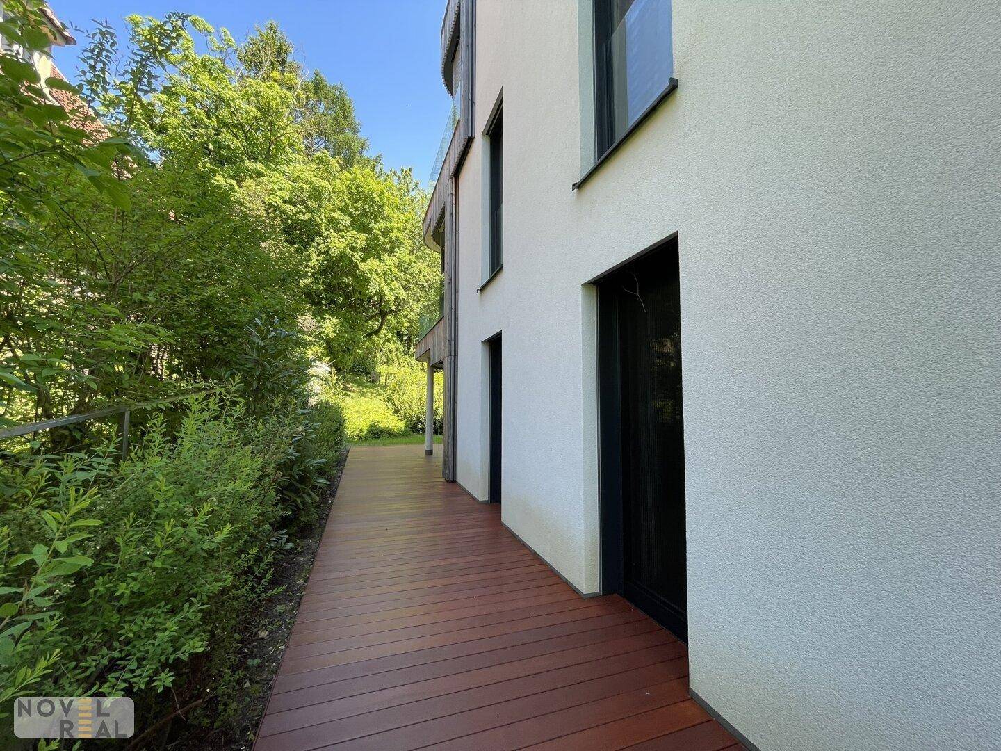 Seitlicher Zugang zum Gebäude mit Holzterrasse und üppiger Bepflanzung entlang der Fassade.