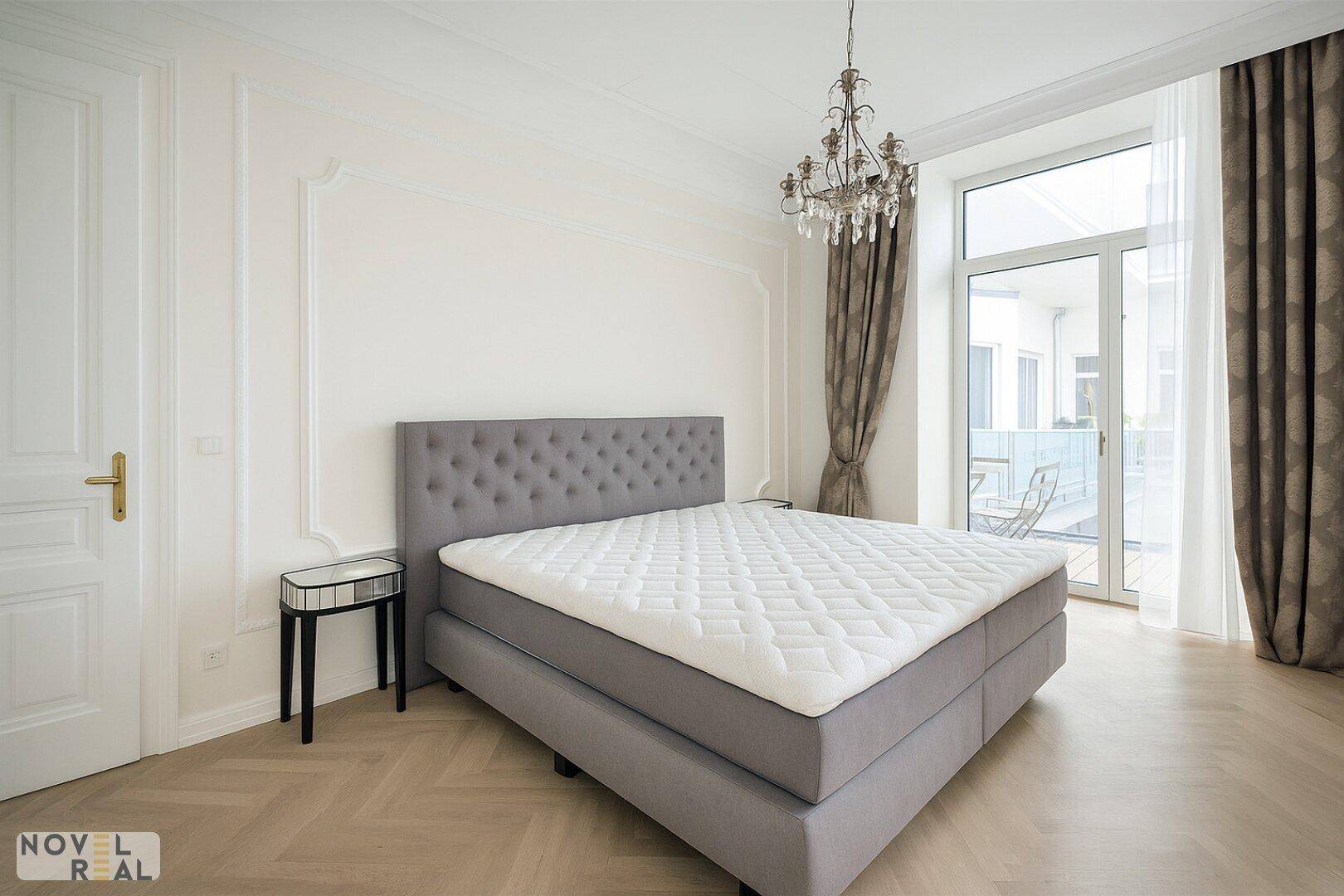 Schlafzimmer mit Fischgrätparkett, Boxspringbett und Zugang zum Balkon.
