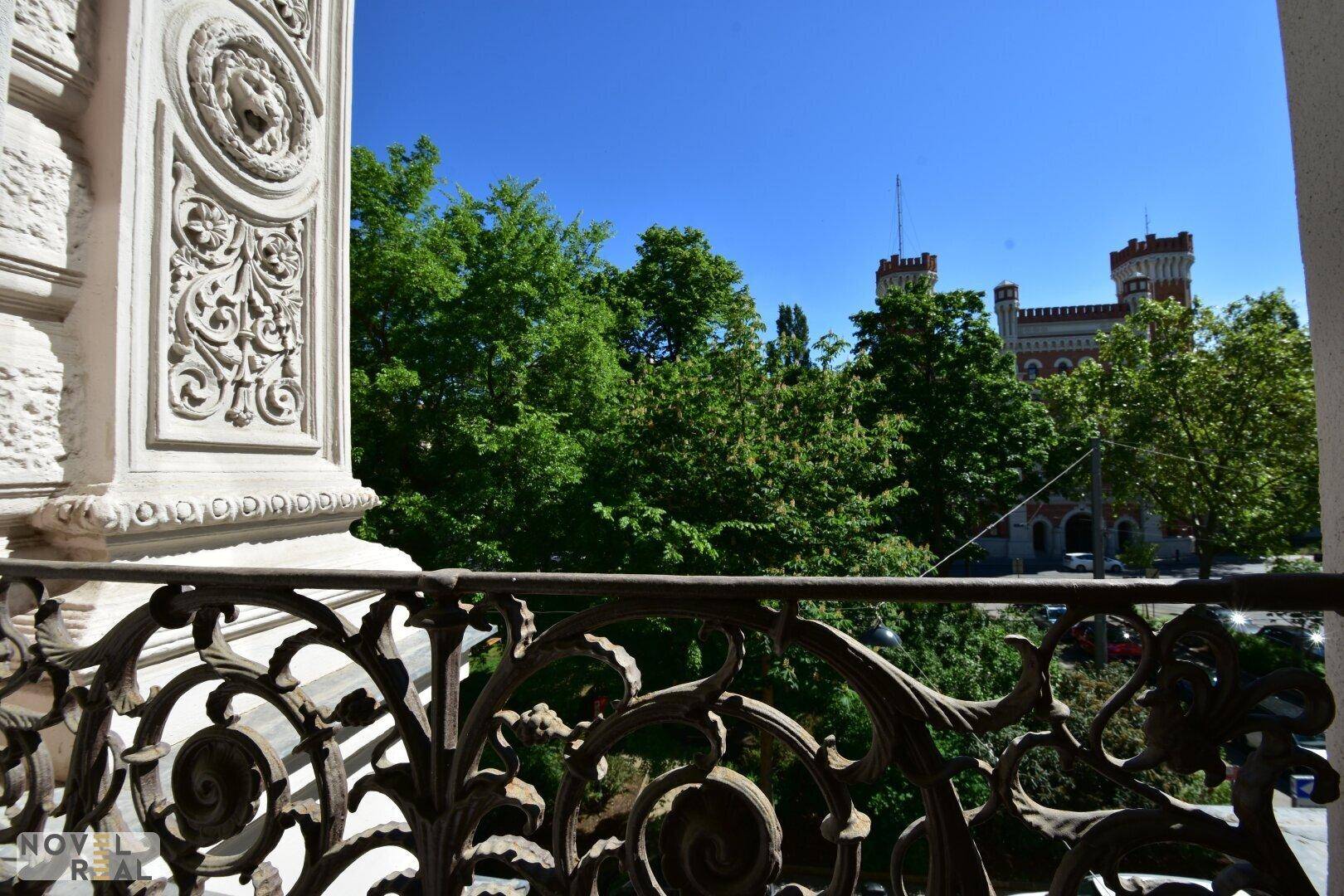Balkon mit schmiedeeisernem Geländer und Blick auf grüne Bäume und ein historisches Gebäude.
