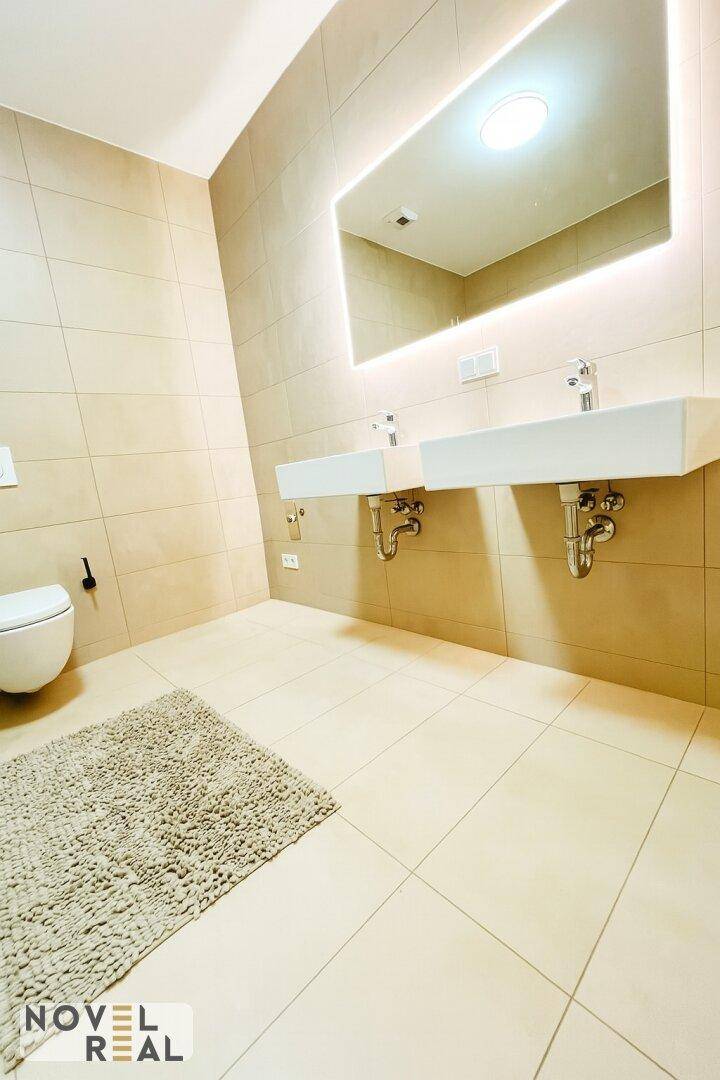 Modernes Badezimmer mit zwei Waschbecken, beleuchtetem Spiegel und beigen Fliesen.