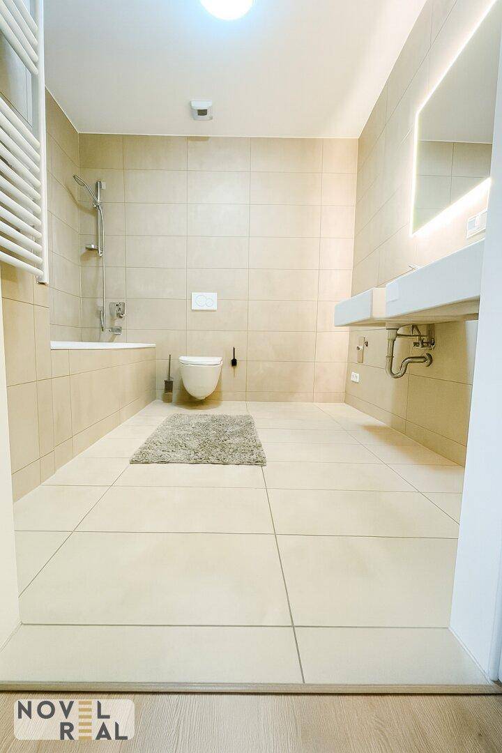 Modernes Badezimmer mit beigen Fliesen, wandhängendem WC und Badewanne mit Duschfunktion.