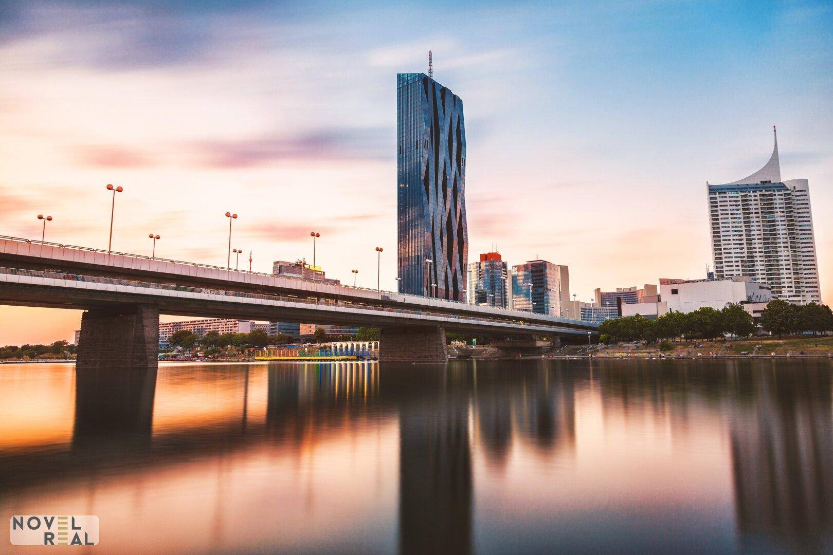 Abendlicher Blick auf die Stadt am Fluss mit Brücke und markanten Hochhäusern bei Sonnenuntergang.