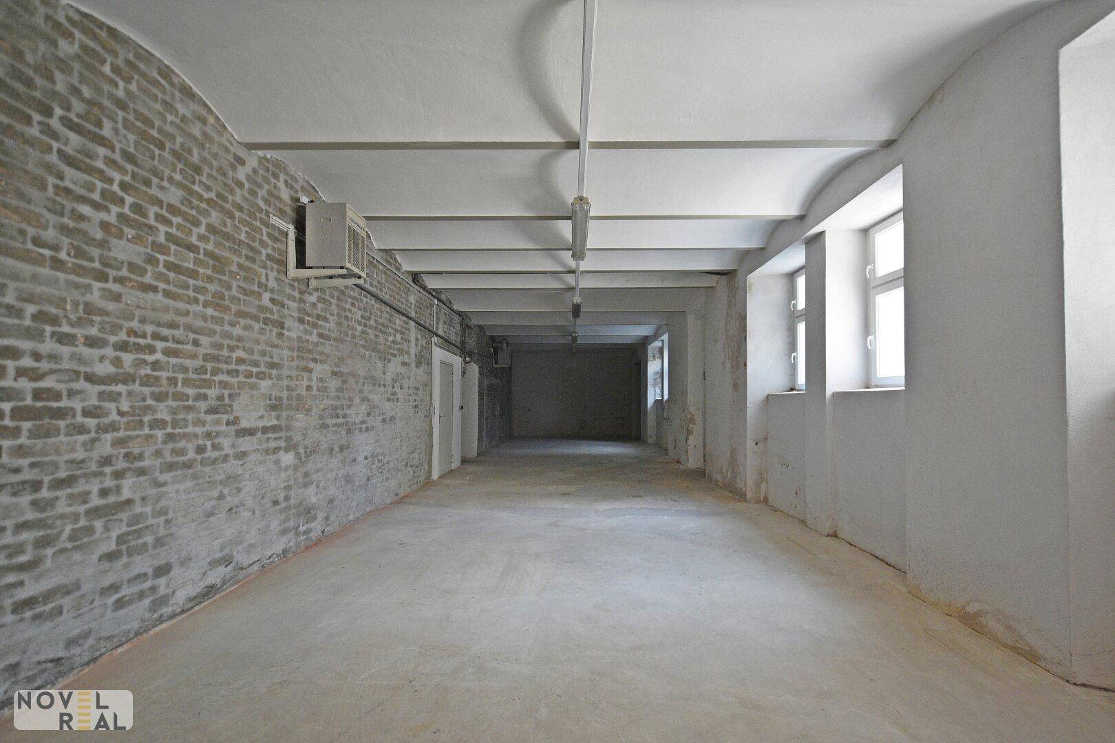 Geräumiger, unmöblierter Raum mit freiliegenden Ziegelwänden, Betonboden und Tageslicht durch Fenster.