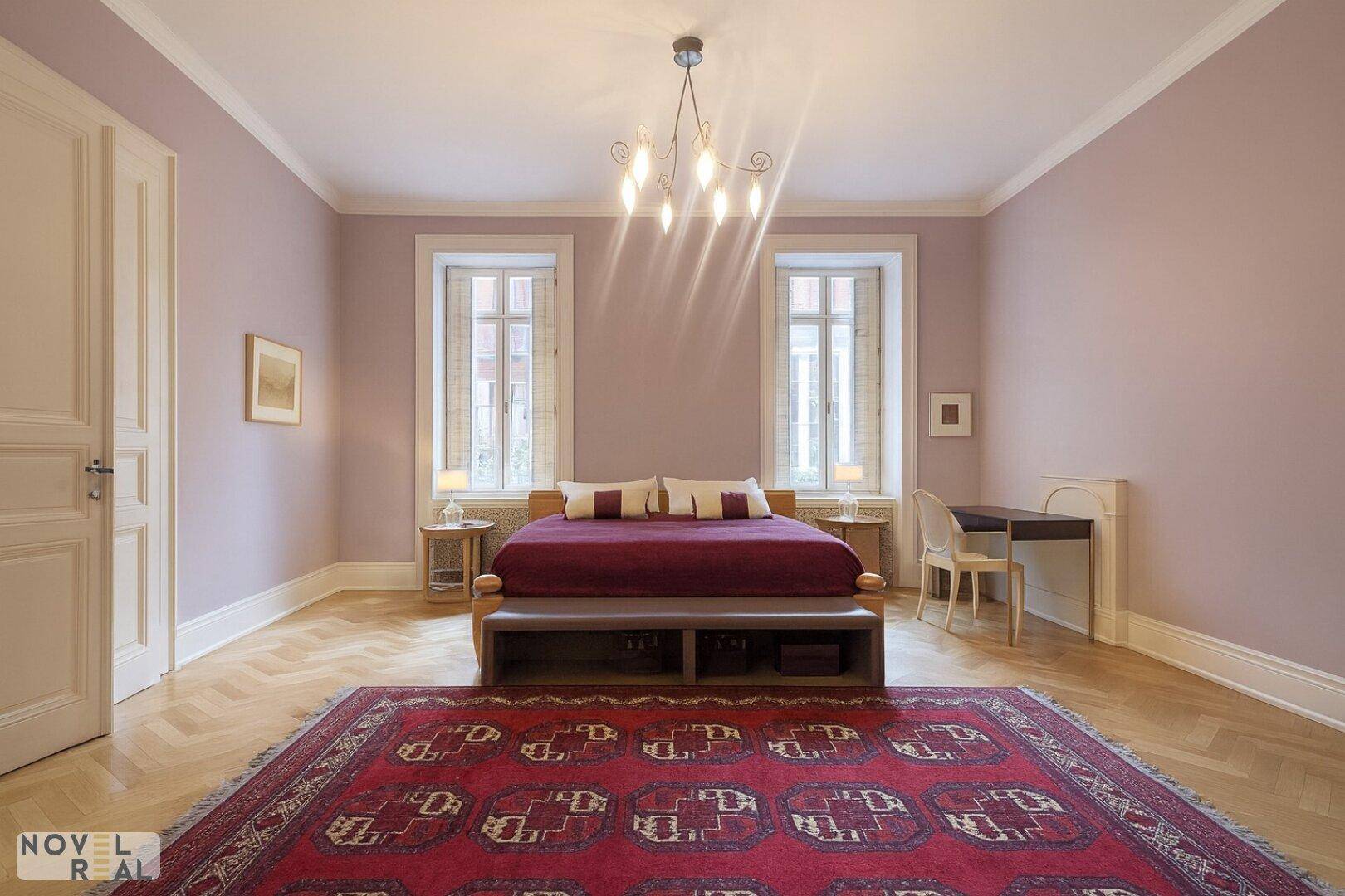Helles Schlafzimmer mit Fischgrätparkett, großem Teppich und hohen Decken.