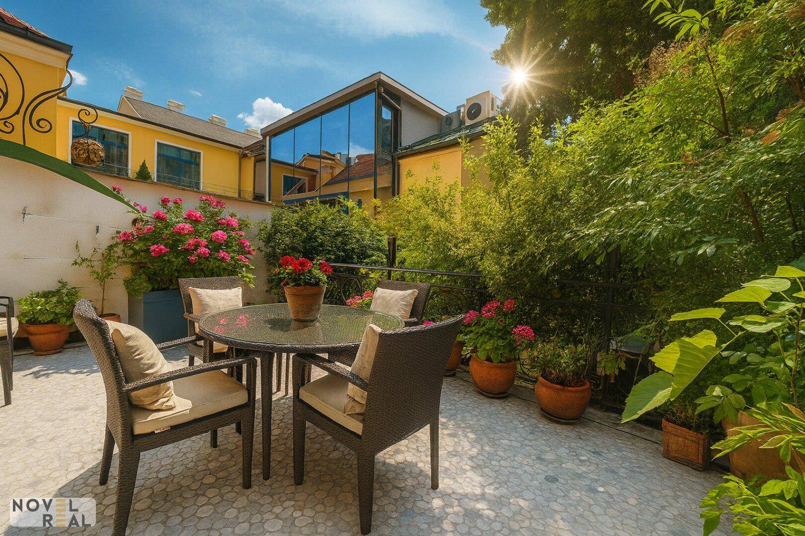 Sonniger Terrassenbereich mit Gartenmöbeln und üppiger Bepflanzung, umgeben von Gebäuden.