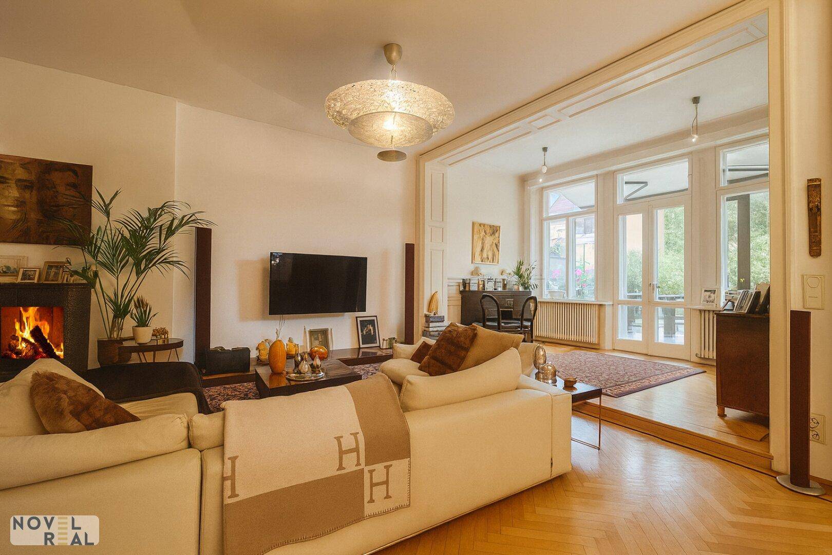 Gemütliches Wohnzimmer mit großem Sofa, Kamin und Zugang zu einem weiteren Raum mit großen Fenstern.