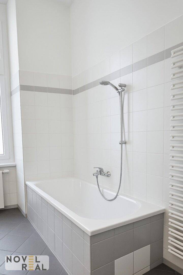 Badewanne mit Handbrause und weiß-grauen Fliesen in einem hellen Badezimmer.