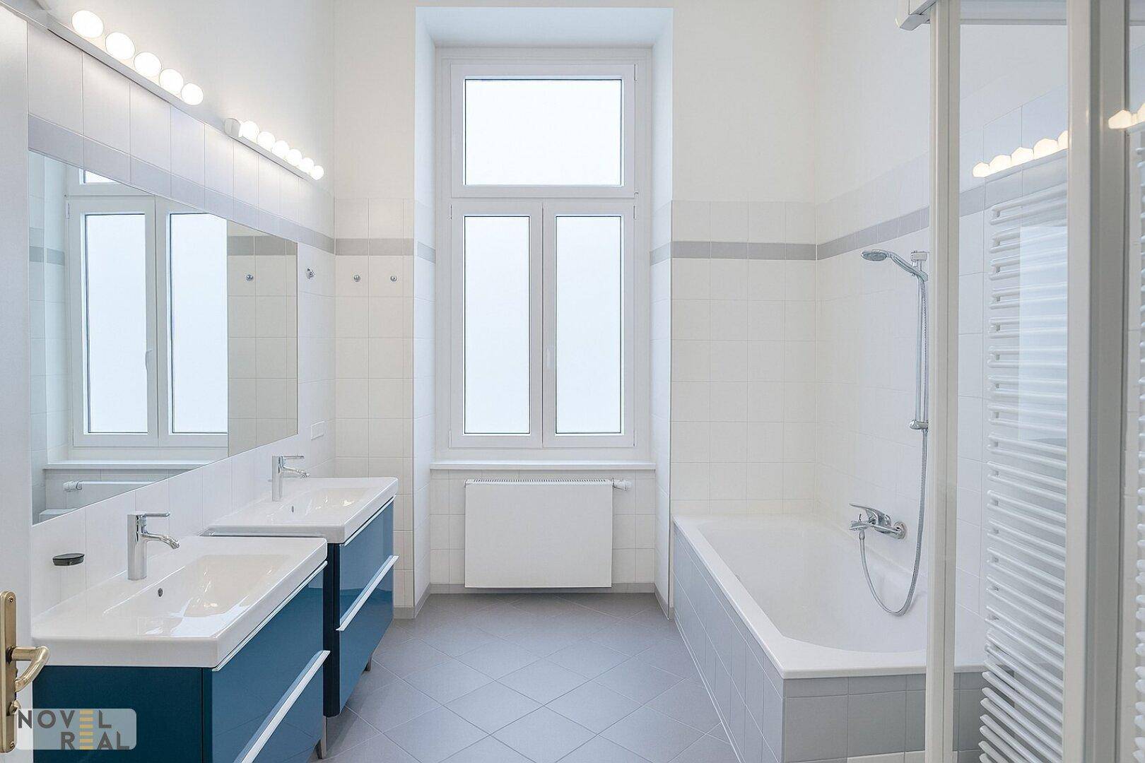Helles Badezimmer mit Doppelwaschbecken, Badewanne, Duschkabine und Fenster.