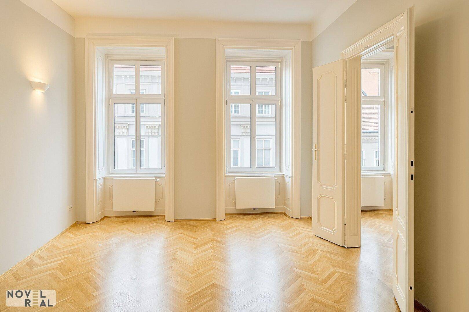 Geräumiges Zimmer mit Fischgrätparkett, hohen Fenstern und klassischer Flügeltür.