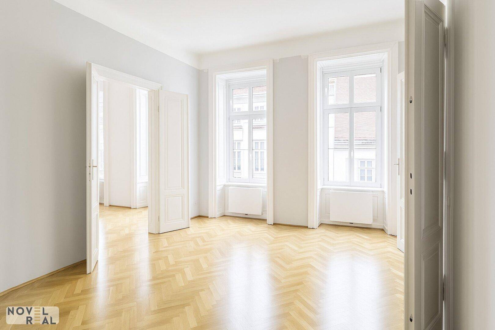 Heller Raum mit Fischgrätparkett und hohen Fenstern, der in weitere Zimmer führt.