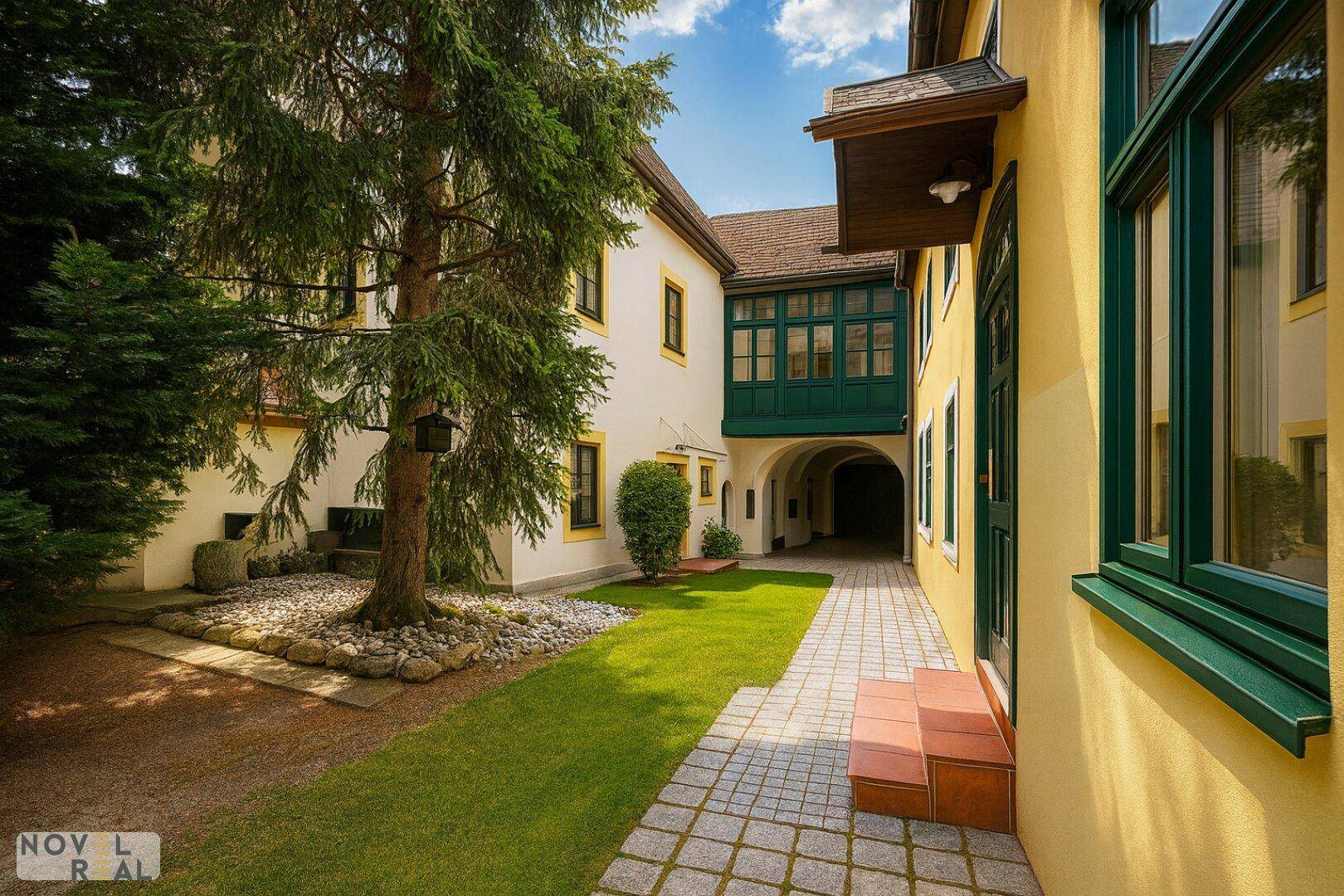 Gepflasterter Innenhof mit altem Baumbestand, einem Durchgang und einer grünen Loggia im Hintergrund.