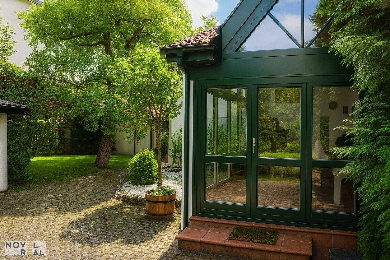Heller Wintergarten mit Glasdach und grünen Rahmen, umgeben von einem gepflegten Garten und Bäumen.