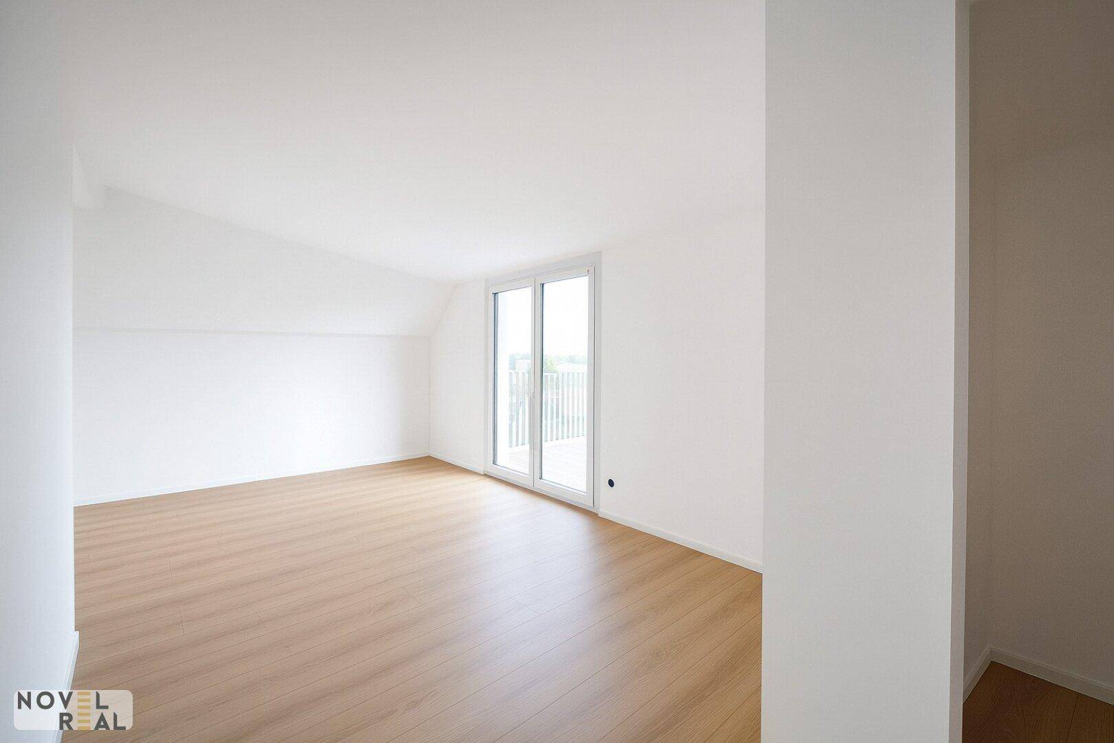 Geräumiges, helles Zimmer mit Dachschräge, Holzboden und Zugang zu einem Balkon oder einer Terrasse.