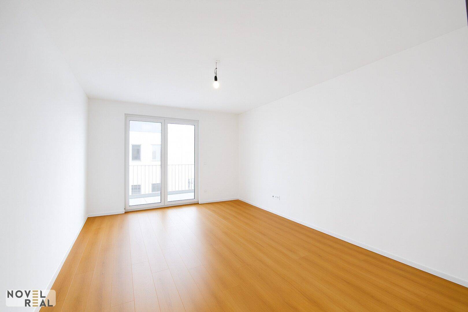 Leerer Raum mit hellem Holzboden und großen Fenstern, die Zugang zu einem Balkon bieten.