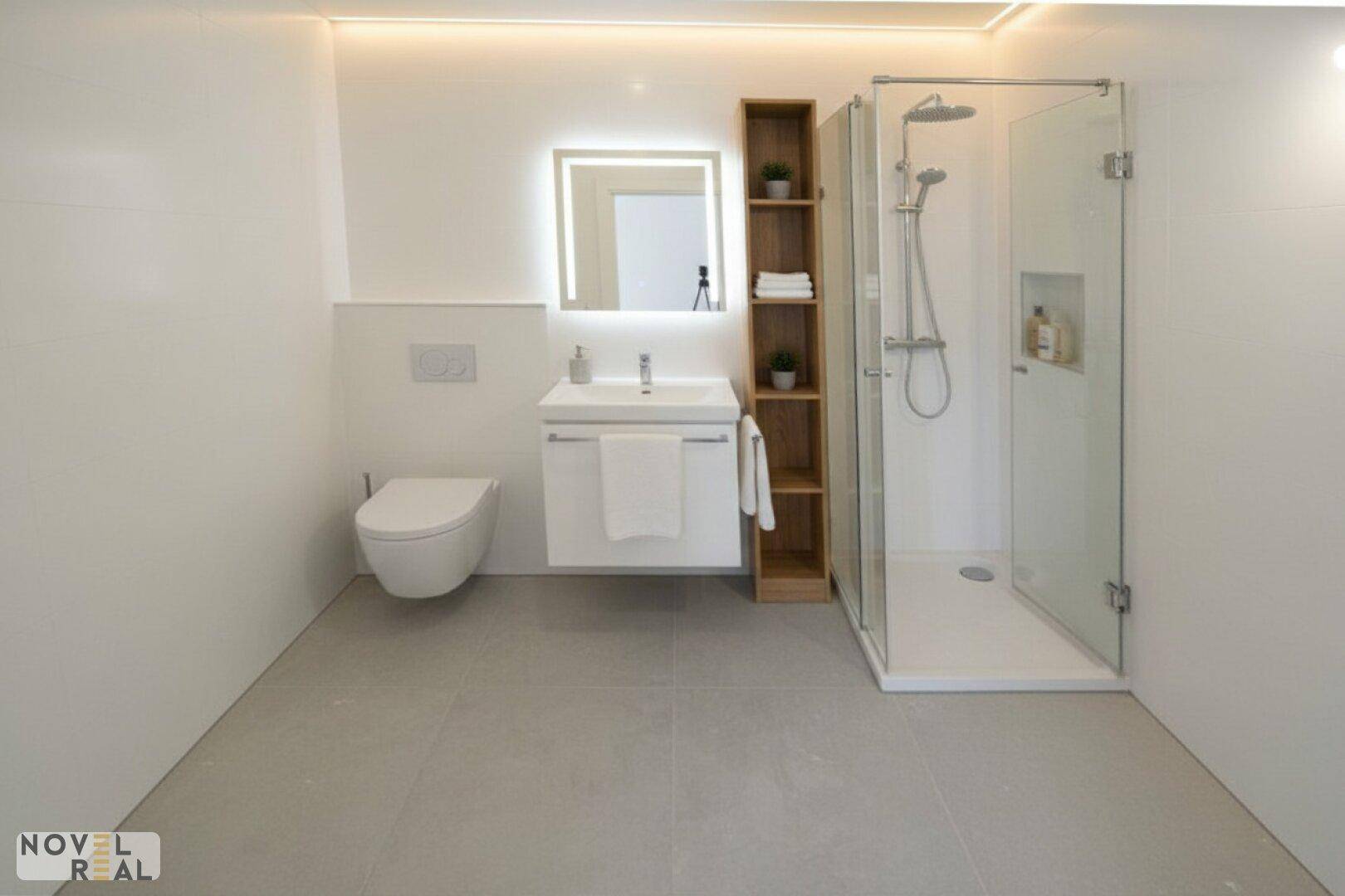 Modernes Badezimmer mit ebenerdiger Dusche, Wand-WC und beleuchtetem Spiegel, in neuwertigem Zustand.
