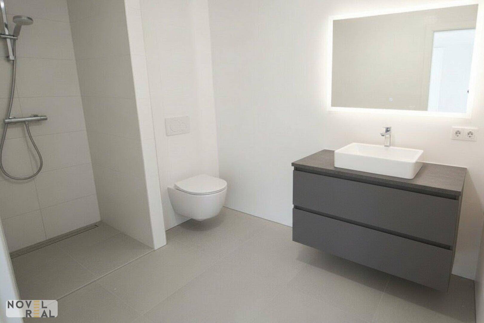 Modernes Badezimmer mit ebenerdiger Dusche, wandhängendem WC und stilvollem Waschtisch mit beleuchtetem Spiegel.