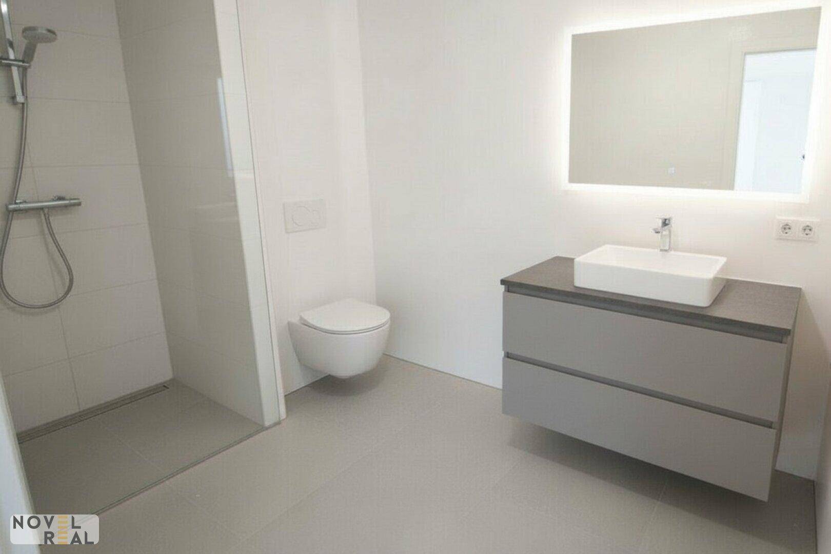 Modernes Badezimmer mit ebenerdiger Dusche, wandhängendem WC und beleuchtetem Spiegel über dem Waschtisch.
