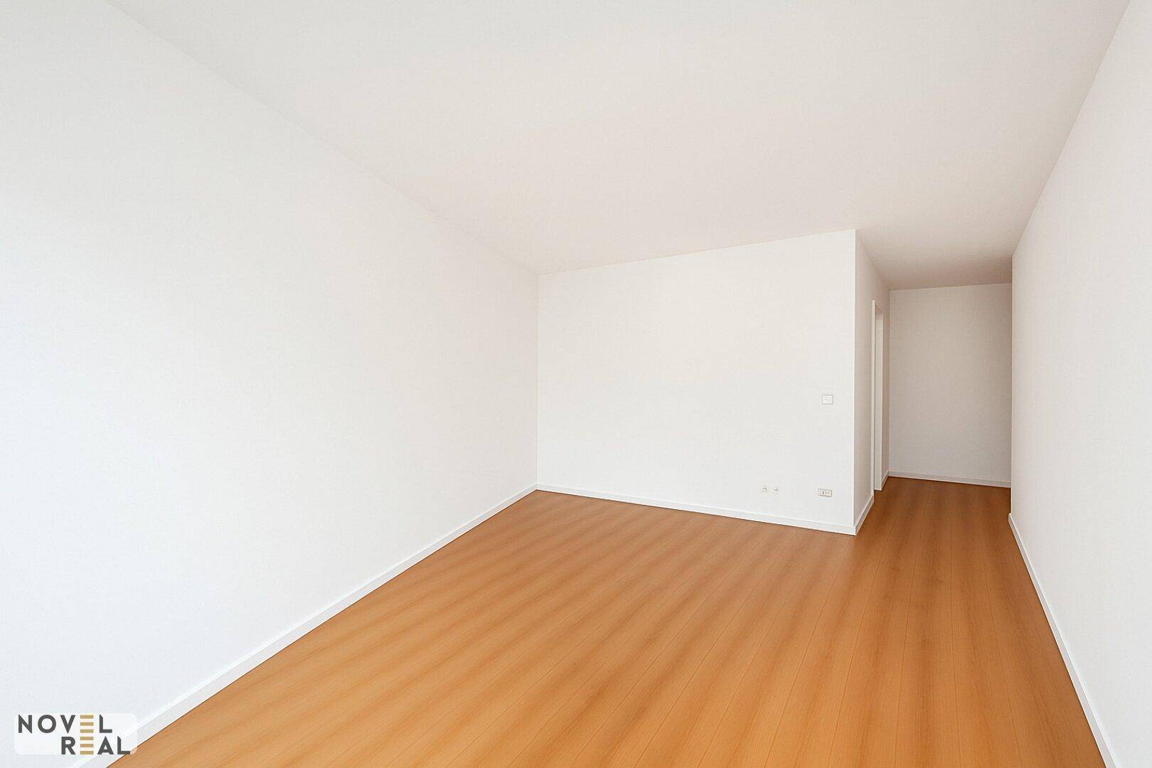 Heller, leerer Raum mit Holzboden und weißen Wänden, der in einen Flur übergeht.