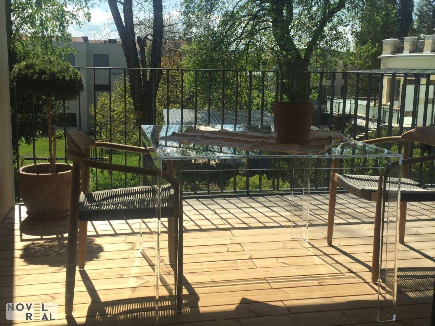 Heller Balkon mit Holzboden, einem modernen Glastisch und Stühlen, umgeben von viel Grün.