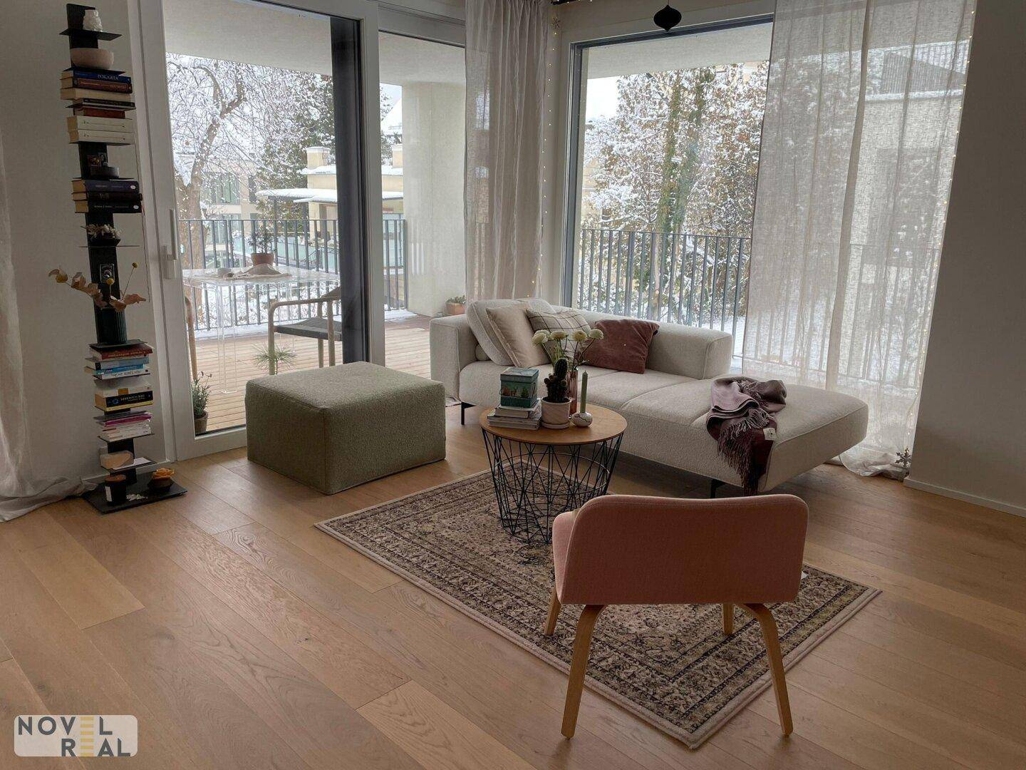 Heller Wohnbereich mit beigem Sofa, Teppich und großen Fenstern mit Blick ins Grüne.