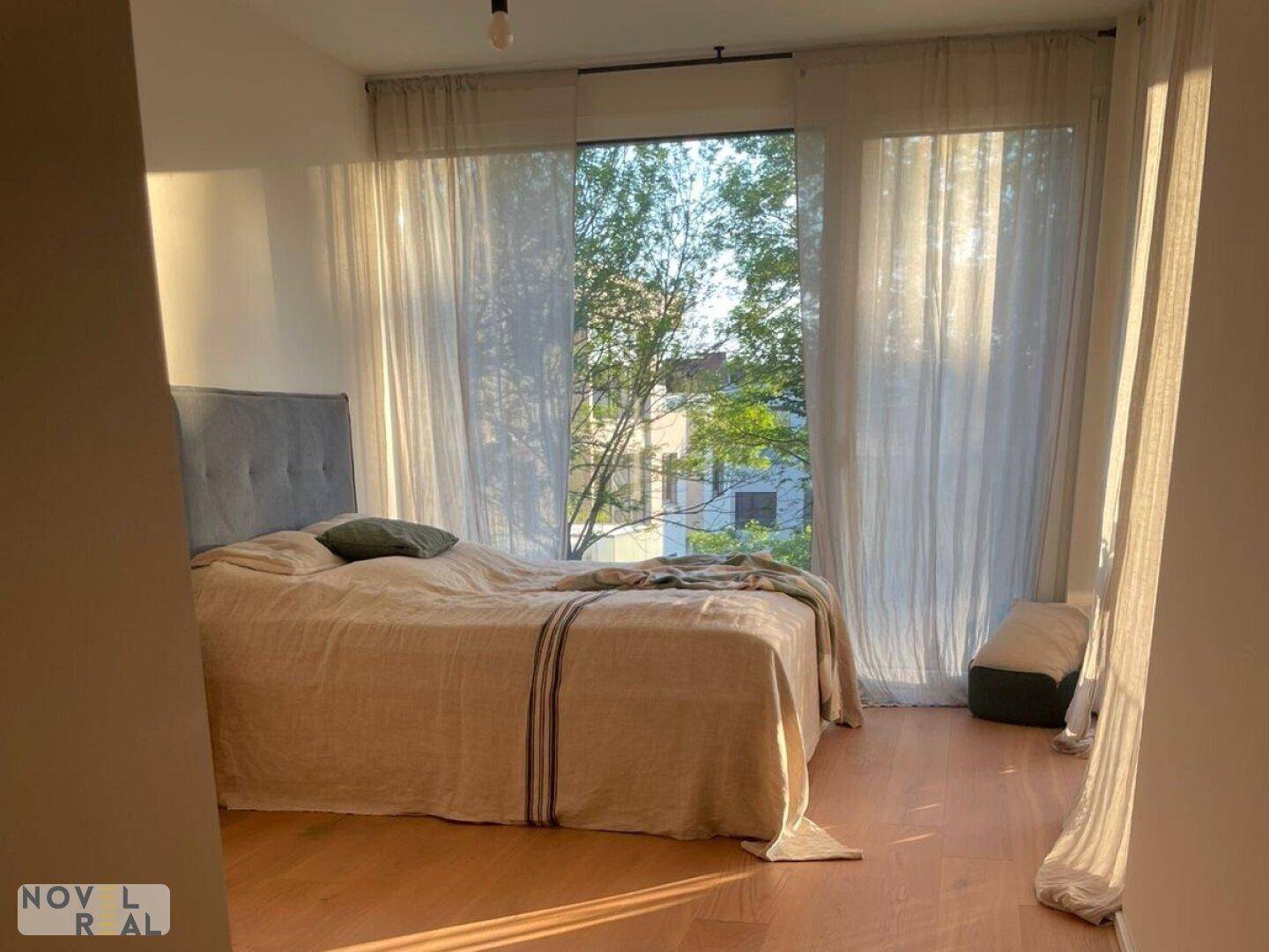 Helles Schlafzimmer mit großem Bett, Holzboden und bodentiefen Fenstern mit viel Tageslicht.