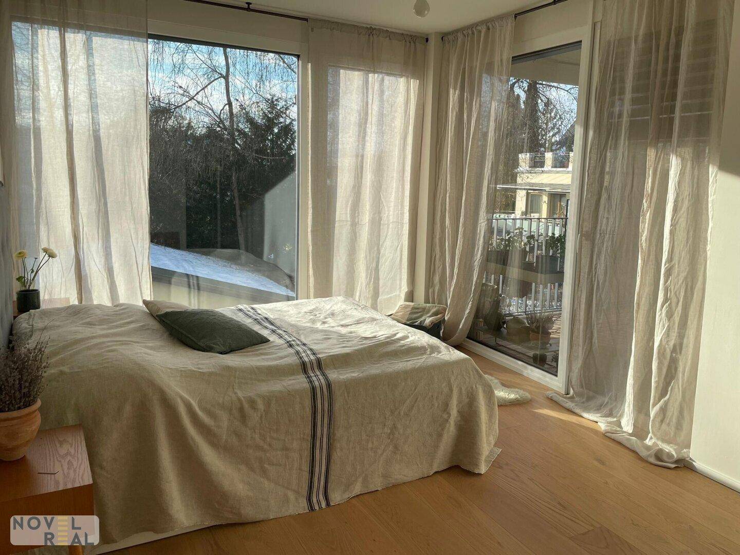 Lichtdurchflutetes Schlafzimmer mit großem Bett, Holzboden und weiten Ausblicken durch die Fenster.