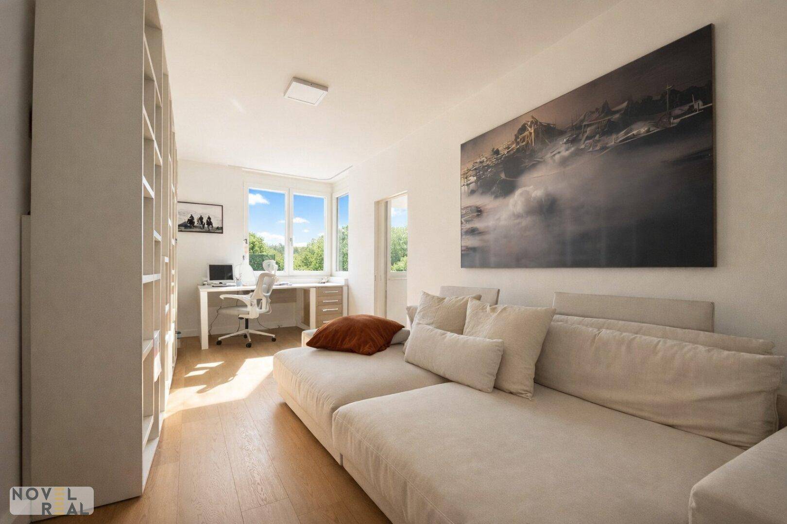 Helles Arbeitszimmer mit Schreibtisch am Fenster, Bücherregal und bequemer Couch.