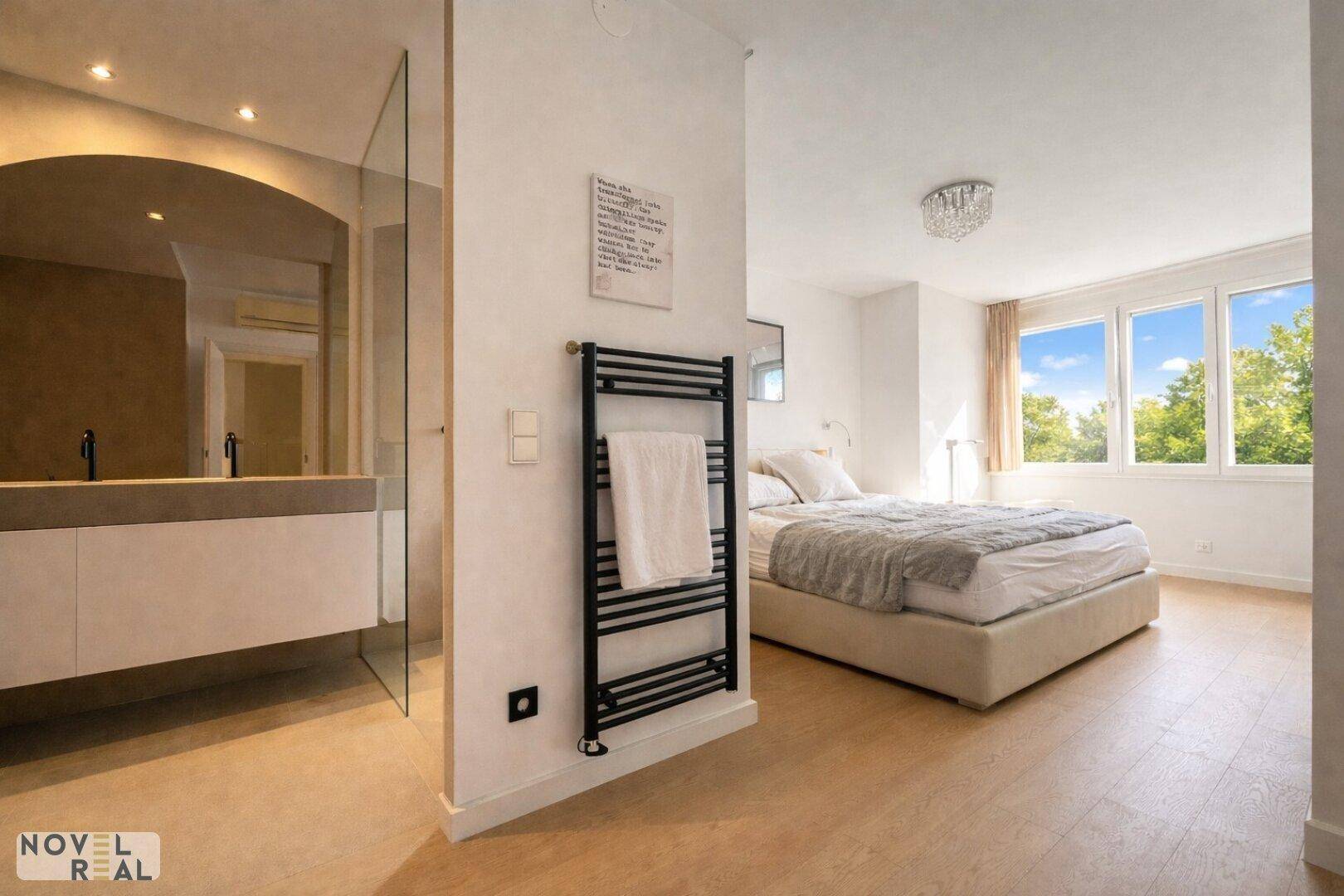 Helles Schlafzimmer mit Holzboden, großen Fenstern und offenem Zugang zum Badezimmer.