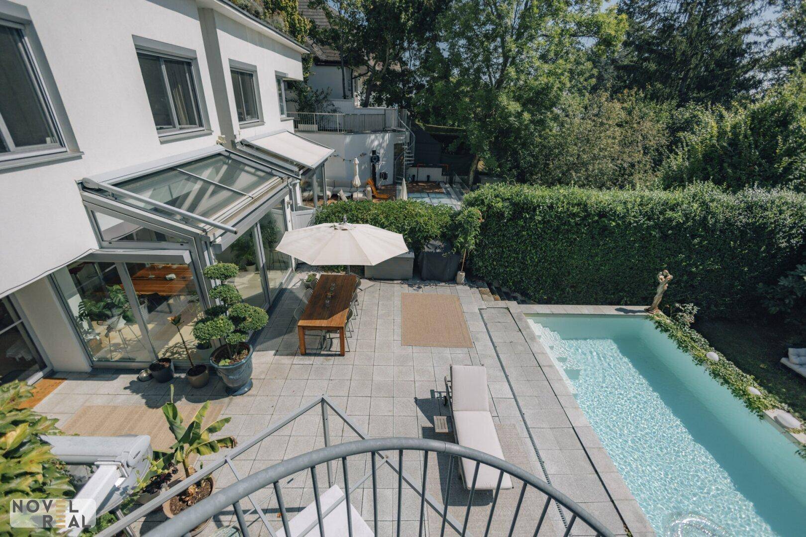 Terrasse mit Essbereich, Sonnenliege und Pool, umgeben von grünen Hecken und Bäumen.