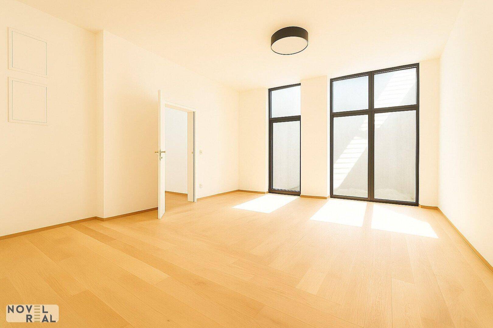 Geräumiges, helles Zimmer mit Holzfußboden und großen Fenstern, die viel Tageslicht hereinlassen.